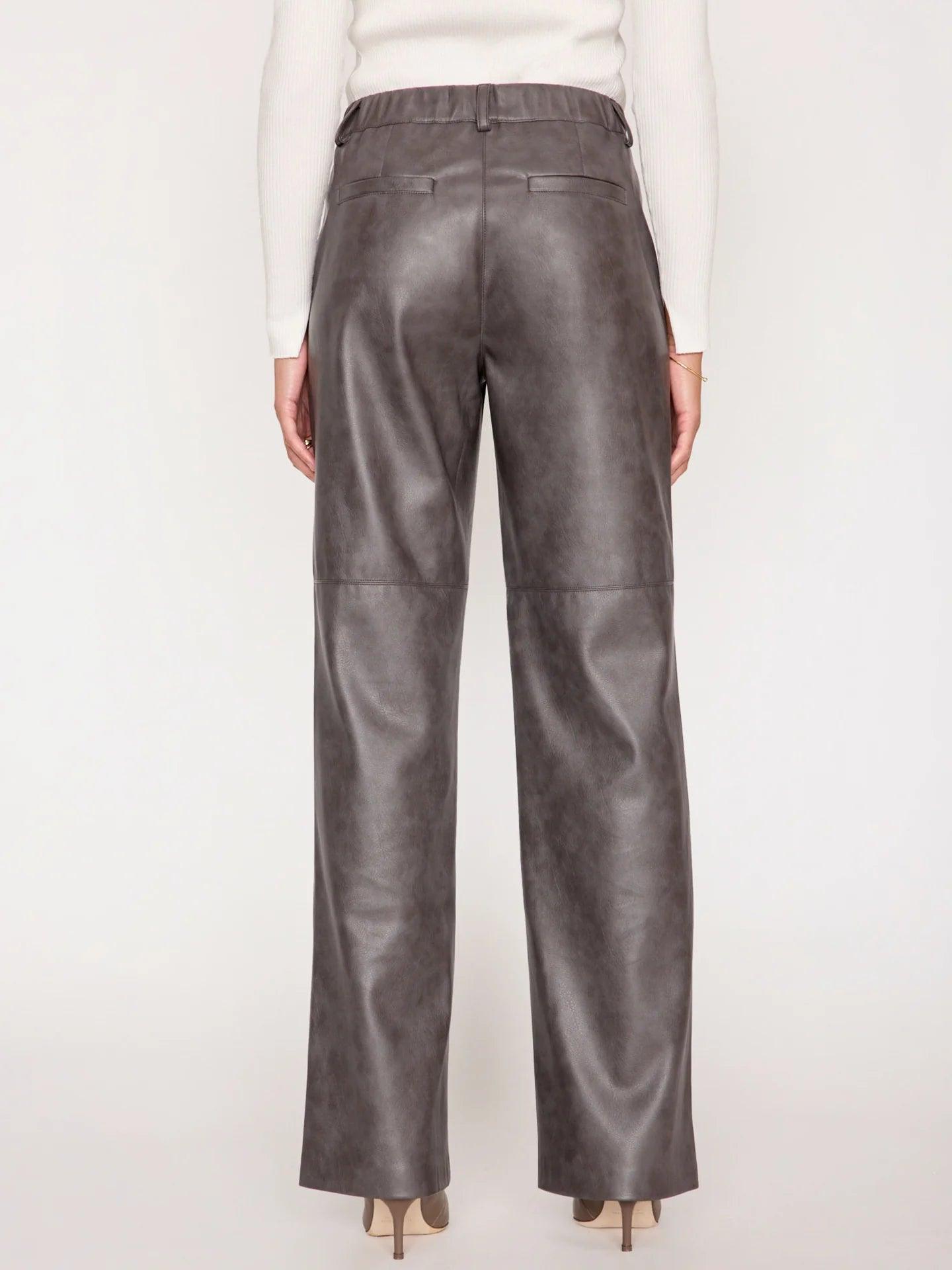 A person stands facing away from the camera, showcasing a pair of high-rise, relaxed-fit Brochu Walker Namari Vegan Leather Pants. They are wearing a white long-sleeve top and beige heeled shoes. The image emphasizes the fit and texture of these luxurious pants by Brochu Walker.