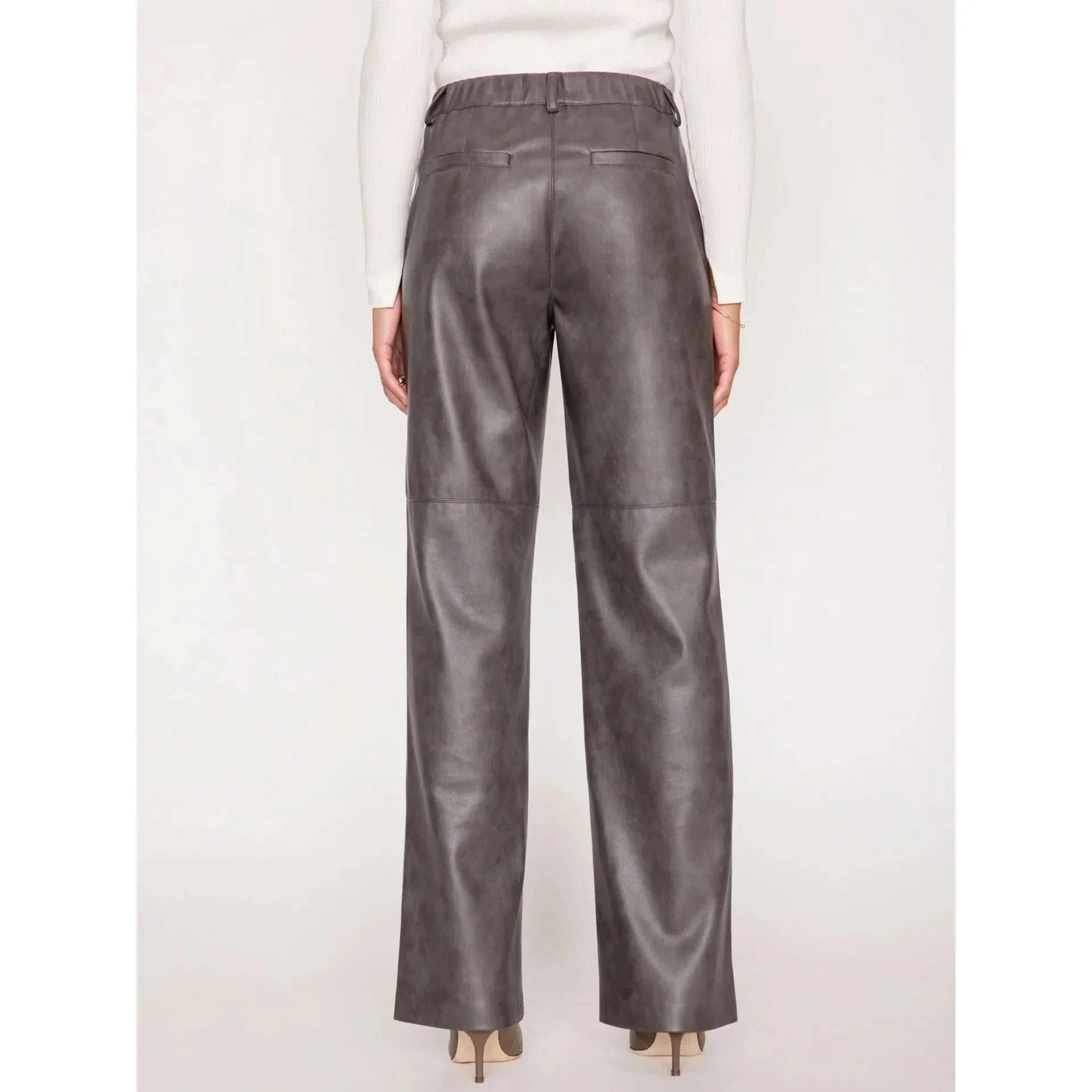 A person stands facing away from the camera, showcasing a pair of high-rise, relaxed-fit Brochu Walker Namari Vegan Leather Pants. They are wearing a white long-sleeve top and beige heeled shoes. The image emphasizes the fit and texture of these luxurious pants by Brochu Walker.