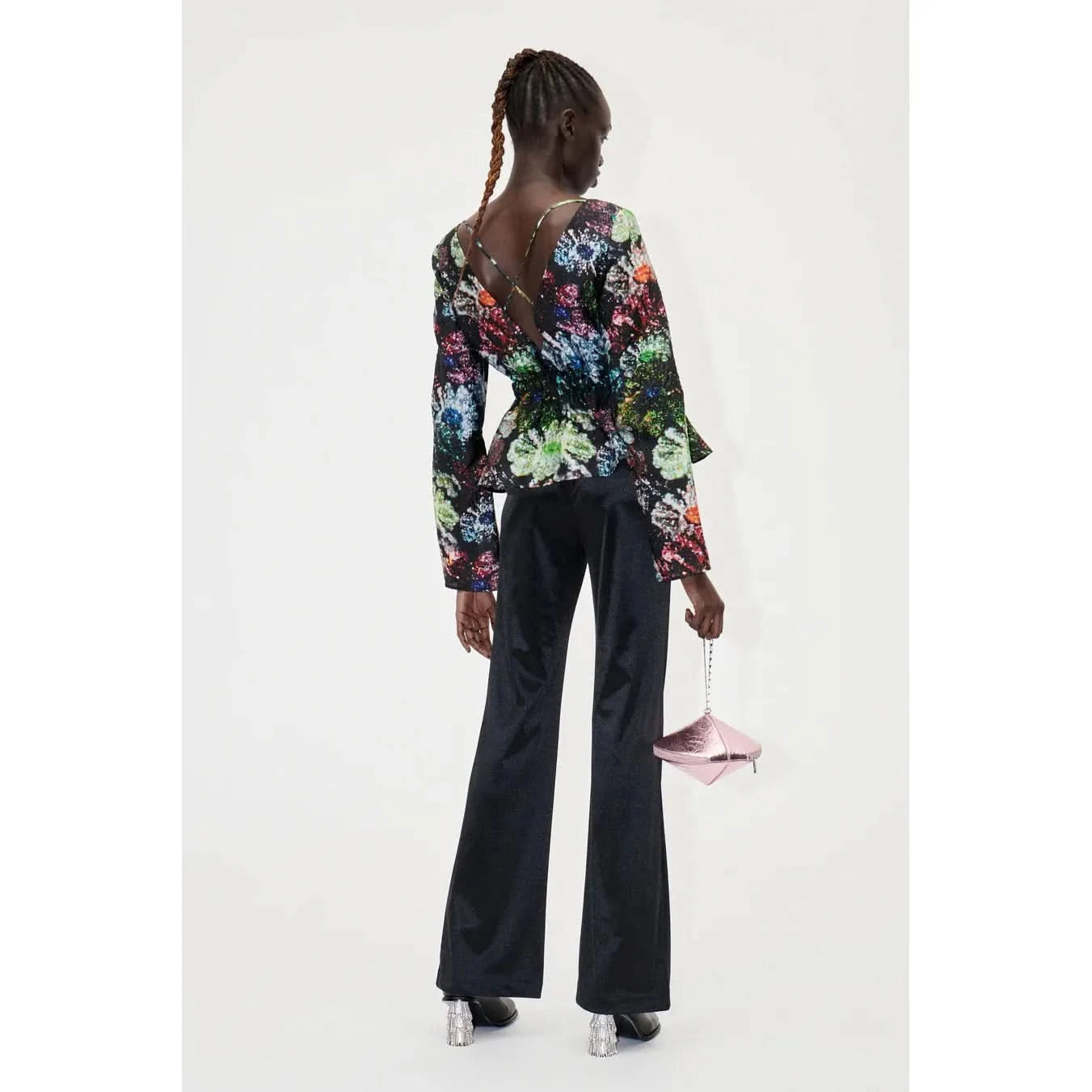 A person with braided hair is standing and facing away from the camera. They are wearing the Stine Goya Nathalie Top Glitter Bloom, a long-sleeved, colorful floral top with an open-back crisscross design made from sustainably sourced materials, black flared pants with a smocked waist, and holding a small, light pink bag. They are also wearing silver heeled shoes.