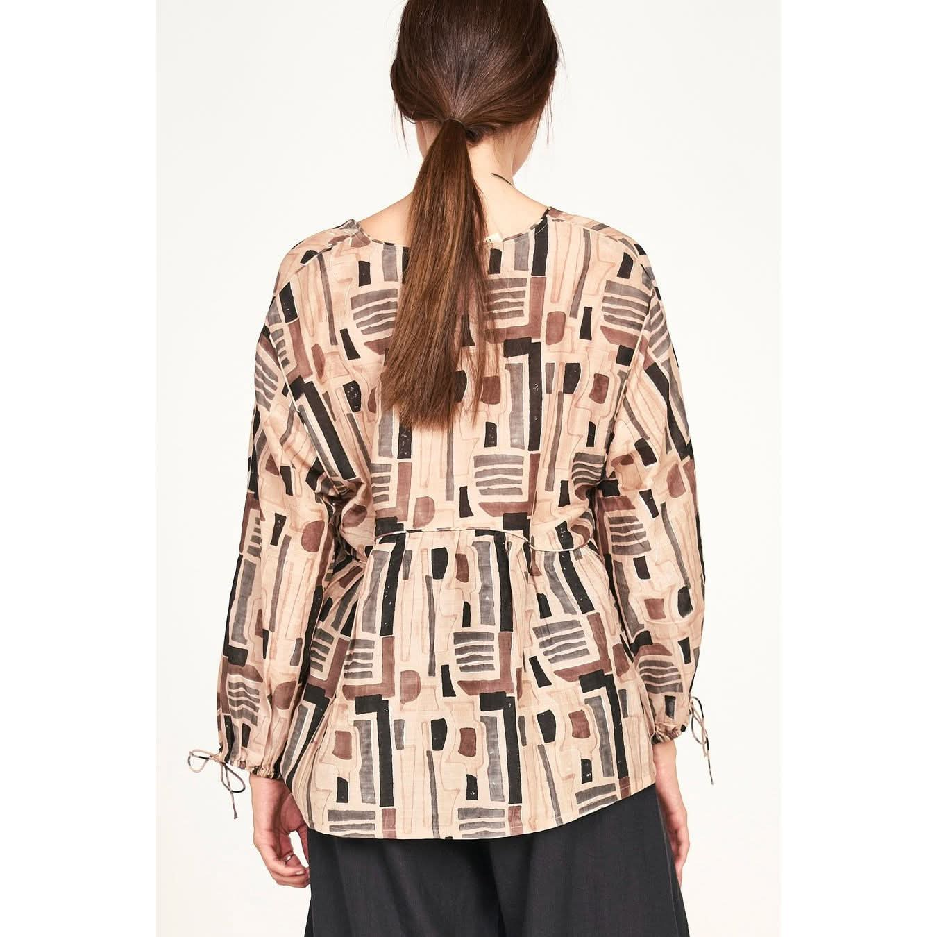 A woman with brown hair in a ponytail wears the Mirth Petaluma Blouse in Sand Labyrinth. This cotton/silk blend top features geometric shapes in black, brown, and gray, boasting long sleeves with drawstring cuffs for an elegant flair from behind. She pairs it seamlessly with dark pants for a chic look.