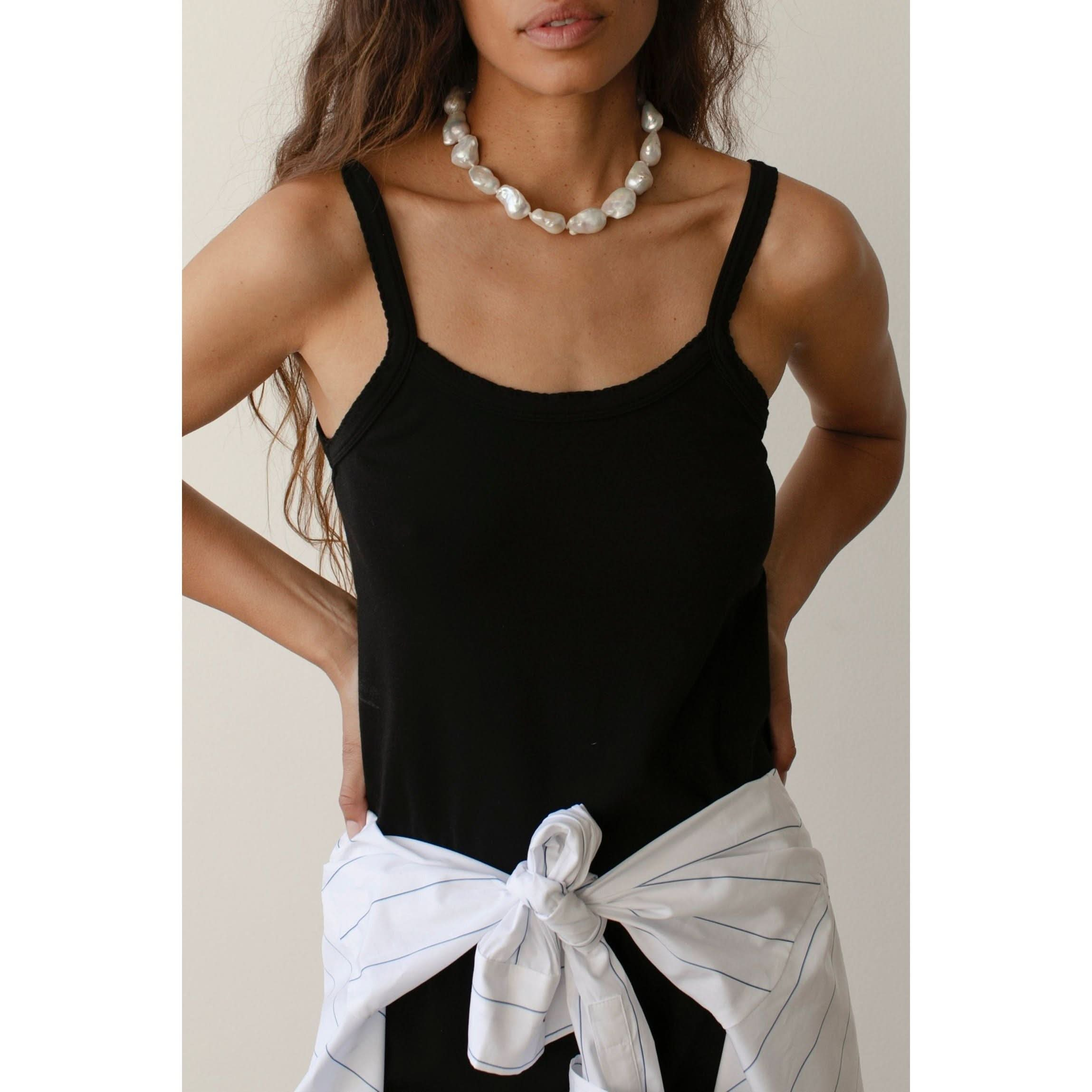 A woman stands wearing a black Donni The Baby Rib Scallop Tank by Donni, featuring scalloped edges and a white necklace made of large beads. She has her hands on her hips, with a white long-sleeve shirt adorned with thin blue stripes tied around her waist. The background is a plain light color.