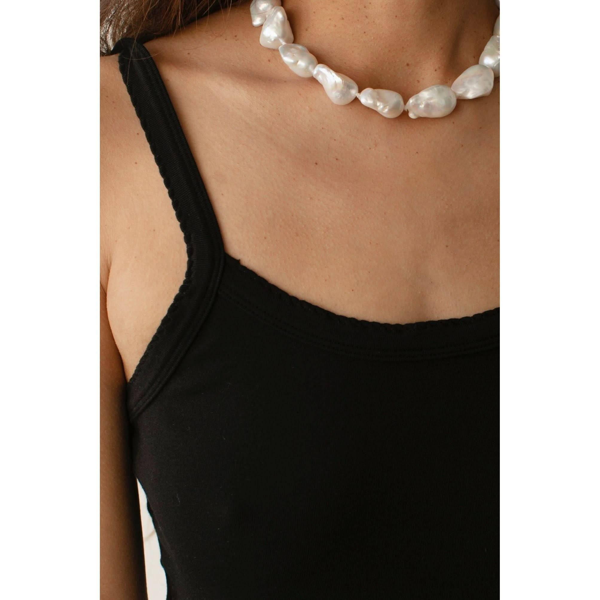 Close-up of a person wearing Donni's The Baby Rib Scallop Tank in black with scalloped edges and a white irregularly-shaped pearl necklace. The image focuses on the neckline, collarbones, and jewelry. The background is not visible.