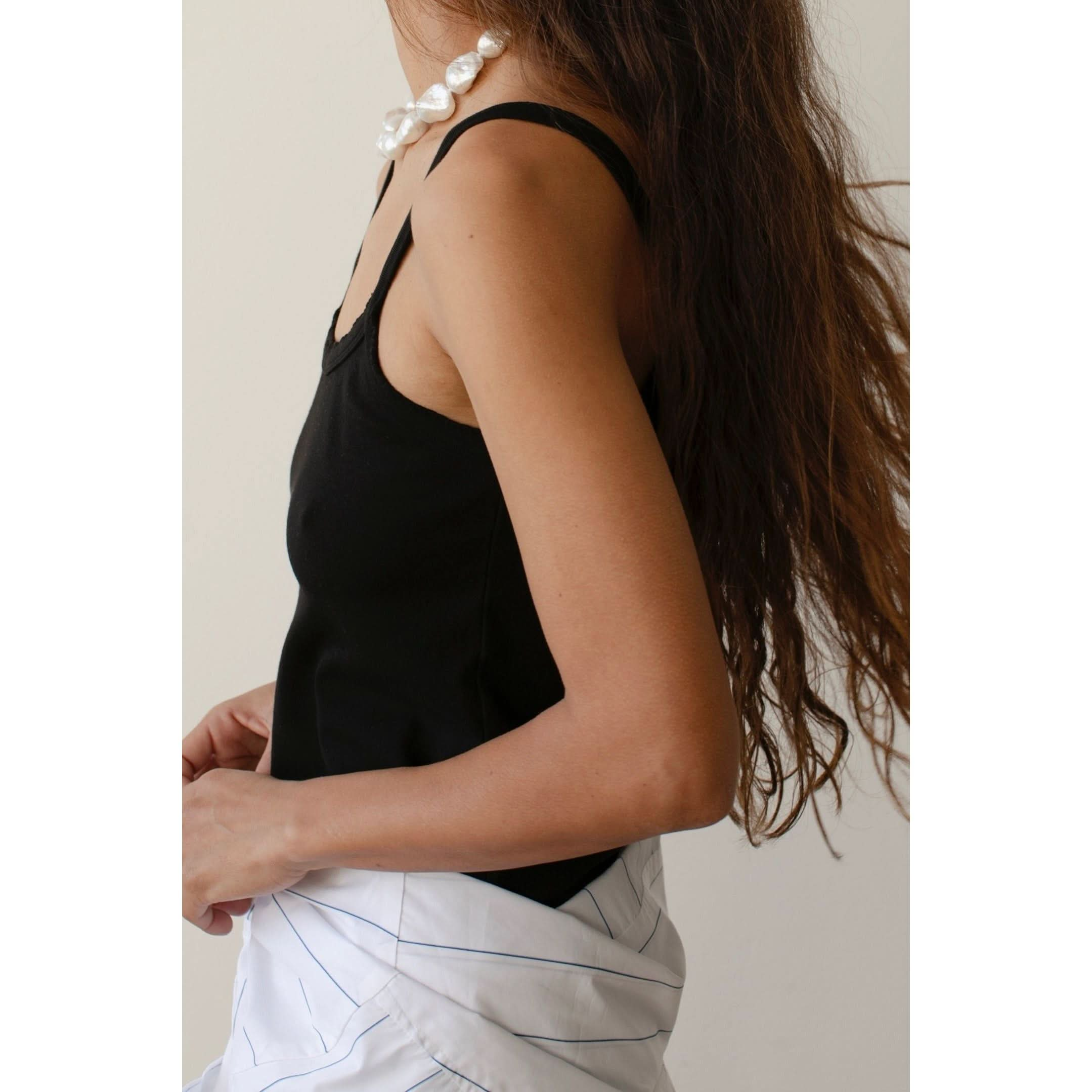 Side view of a person with long, wavy brown hair, wearing the Donni The Baby Rib Scallop Tank with scalloped edges, a necklace adorned with large pearls, and a white garment tied around their waist. The background is plain and light-colored.