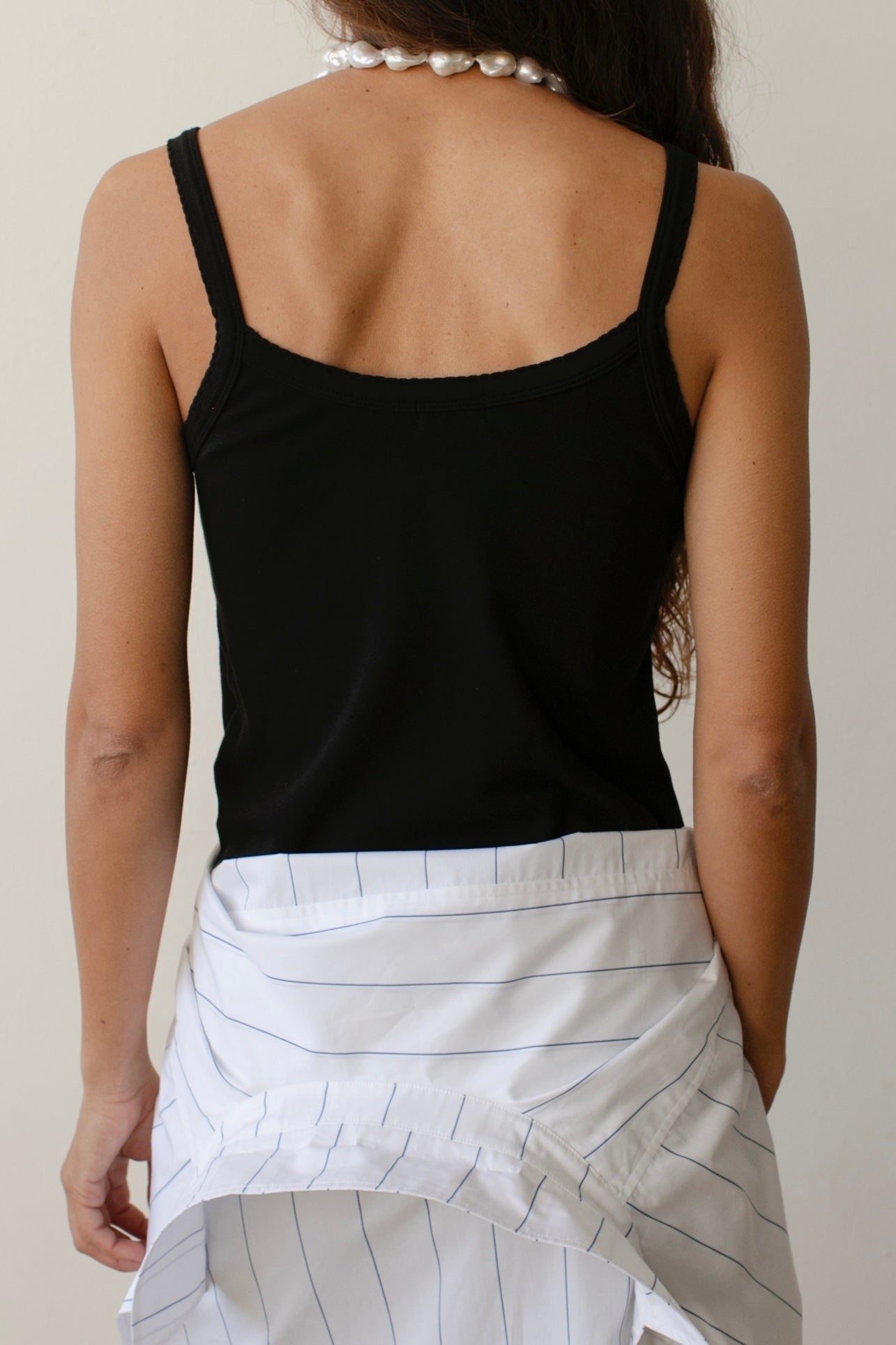 A person with long hair, seen from the back, is wearing Donni's The Baby Rib Scallop Tank in black with a white shirt tied around their waist. The tank features scalloped edges, and the person accessorizes with a pearl necklace.