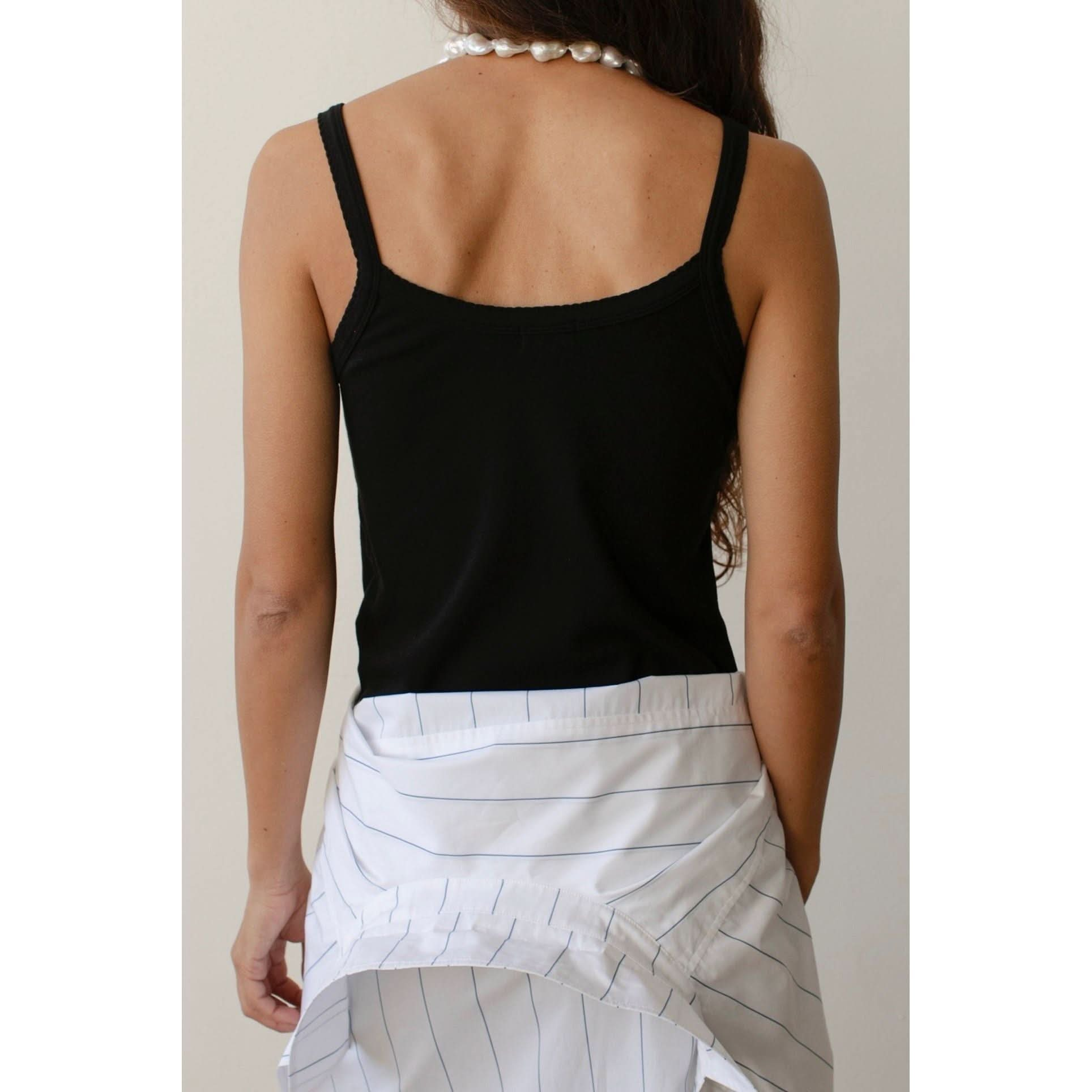 A person with long hair, seen from the back, is wearing Donni's The Baby Rib Scallop Tank in black with a white shirt tied around their waist. The tank features scalloped edges, and the person accessorizes with a pearl necklace.