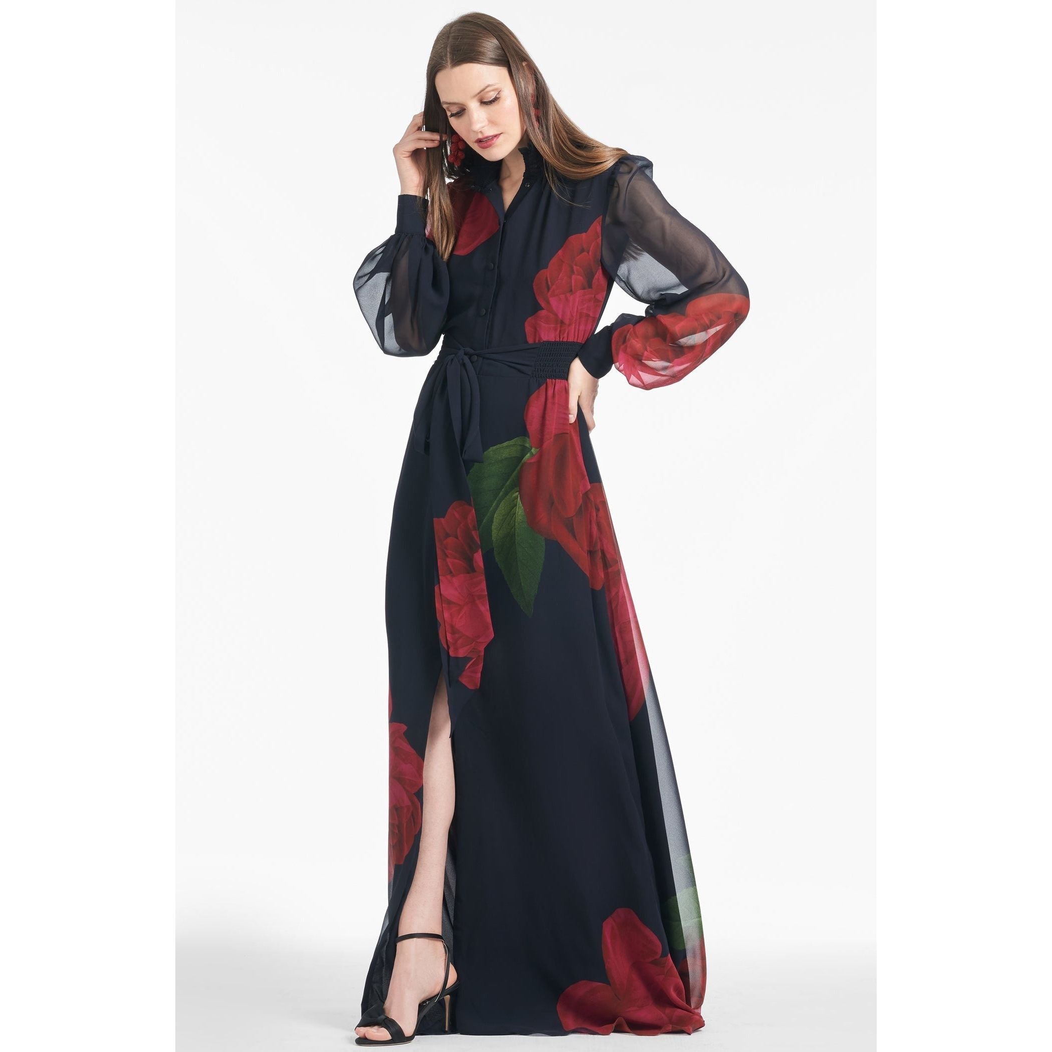 A woman wearing the Sachin & Babi Cassandra Gown in Ruby Rose, featured in a long black dress with prominent red floral designs. The dress is styled with sheer, long sleeves and a waist tie. She is holding her hair with one hand and wearing black high-heeled sandals.