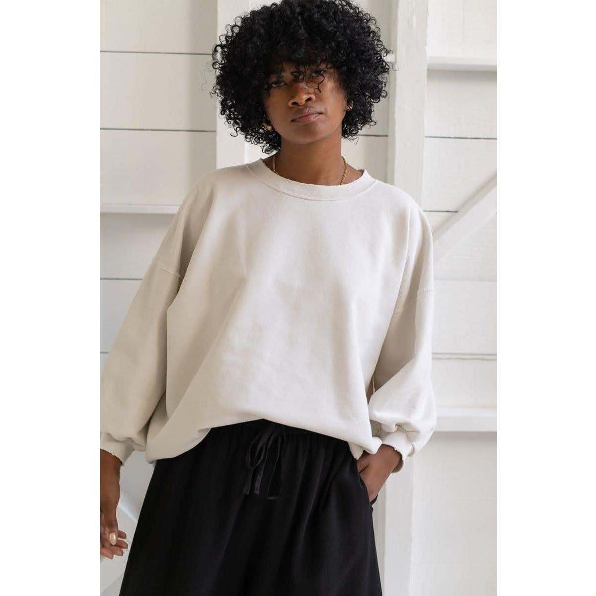 A person with curly hair wears the Rachel Comey Fond Sweatshirt in Dirty White paired with black pants, standing against a white background with shelving. They have a calm expression and one hand tucked into their pocket.