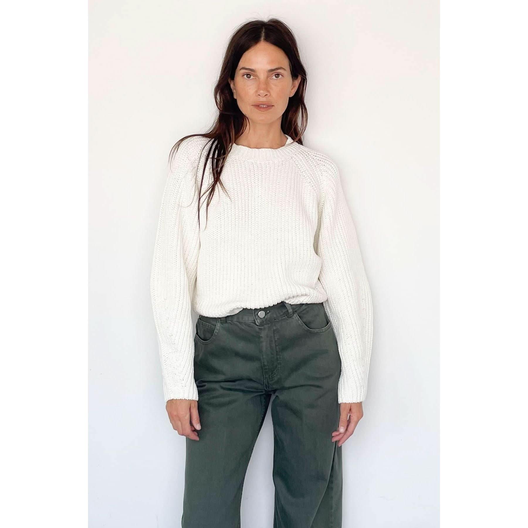 A woman with long hair stands against a simple white background, wearing the Rachel Comey Salla Sweater—a chunky knit piece featuring balloon sleeves—and olive green pants, looking directly at the camera with a neutral expression.