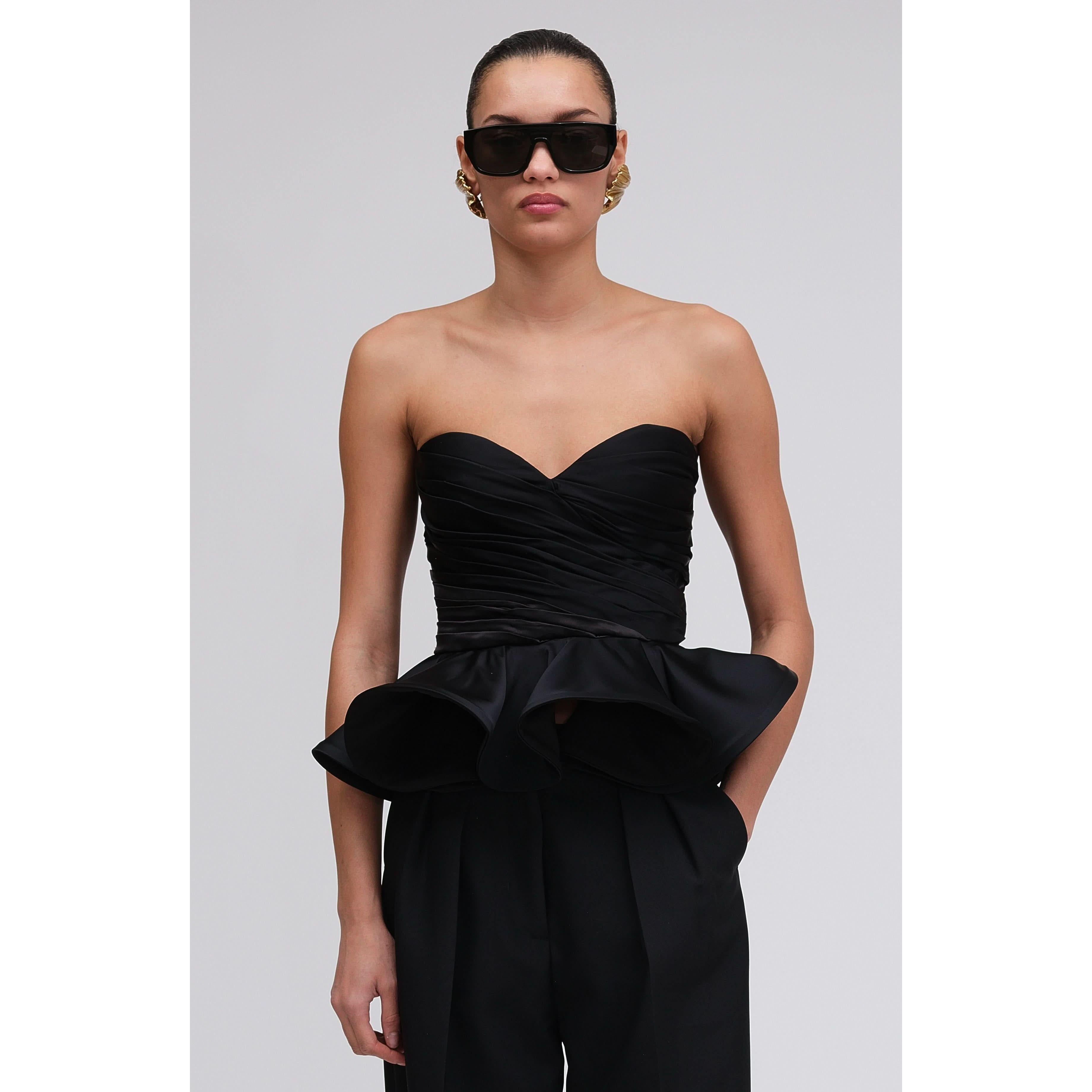 Wearing the Ronny Kobo Tara Top, a chic black strapless piece with ruffled peplum and ruched details, paired with high-waisted black pants instantly channels Fall 2024 style. Hair pulled back elegantly, accessorized with large sunglasses and gold hoop earrings, completes the look against a plain light gray backdrop.