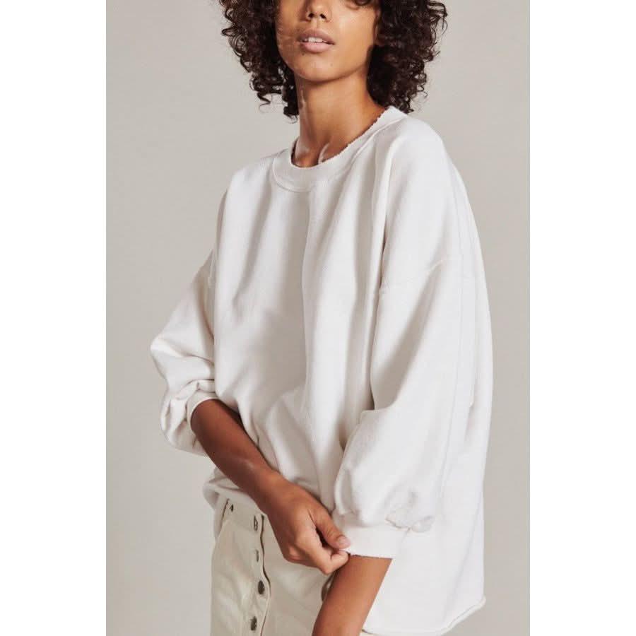 A person with curly hair wears an oversized Rachel Comey Fond Sweatshirt in dirty white long-sleeve cotton and beige pants. They stand against a plain background, gently holding the hem of a sleeve adorned with a subtle ribbed knit pattern.