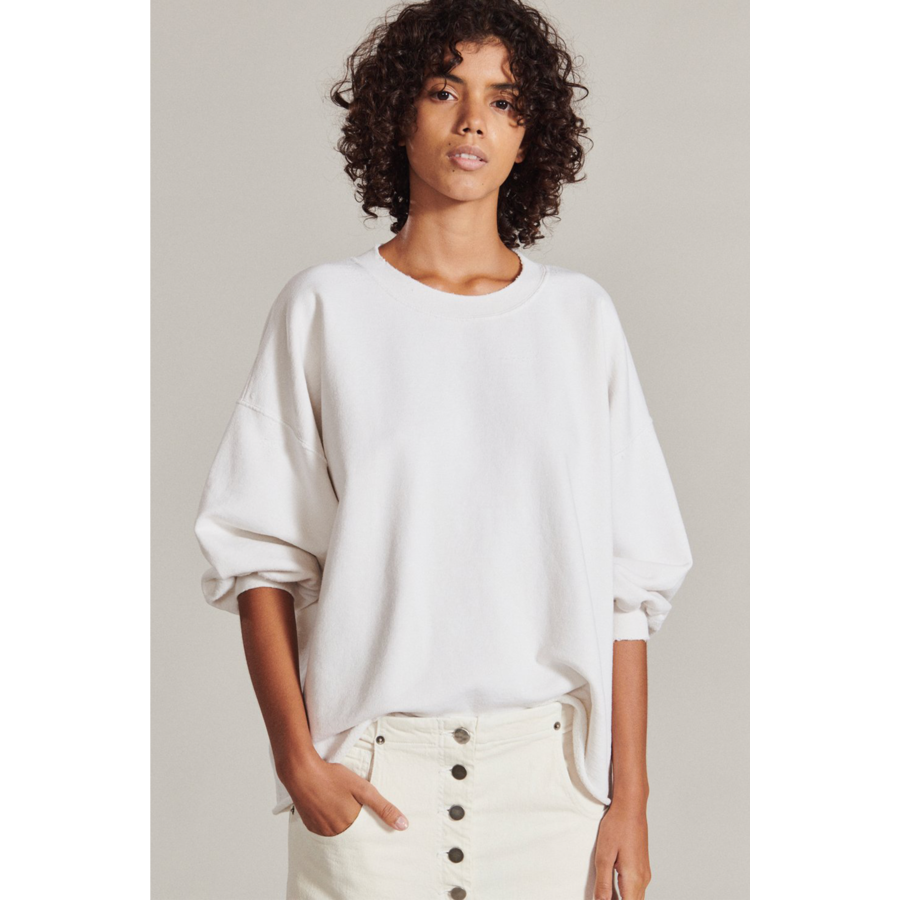 A person with curly hair stands against a plain backdrop wearing a loose-fitting Rachel Comey Fond Sweatshirt in Dirty White and a white skirt with buttons. They have a neutral expression and one hand in their pocket.