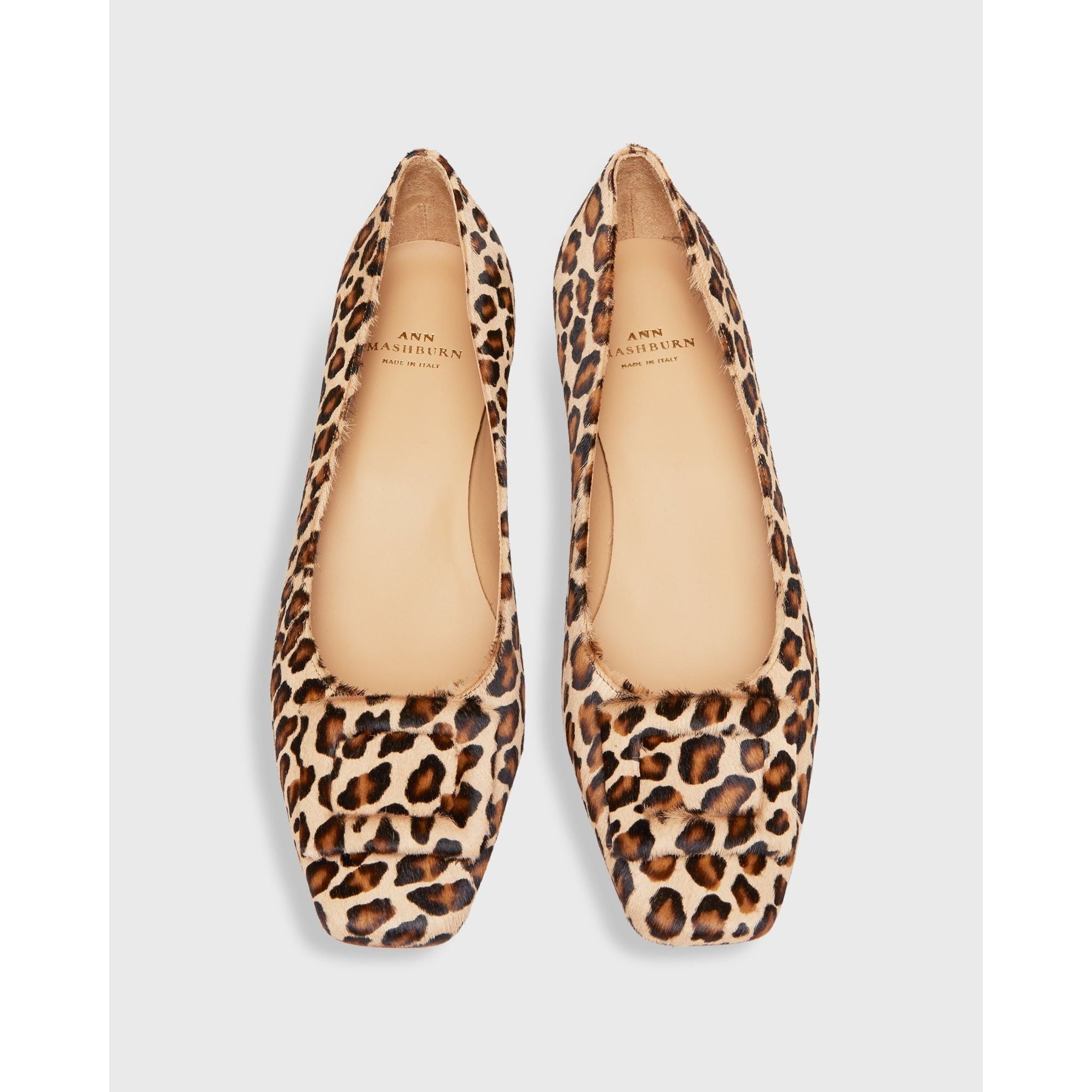 A pair of "Ann Mashburn Buckle Shoe Leopard Calf Hair" women's ballet flats, showcasing leopard print with square toes and beige interiors, display "Ann Mashburn" branding inside. These elegant shoes feature suede leather accents and are positioned side by side on a white background.