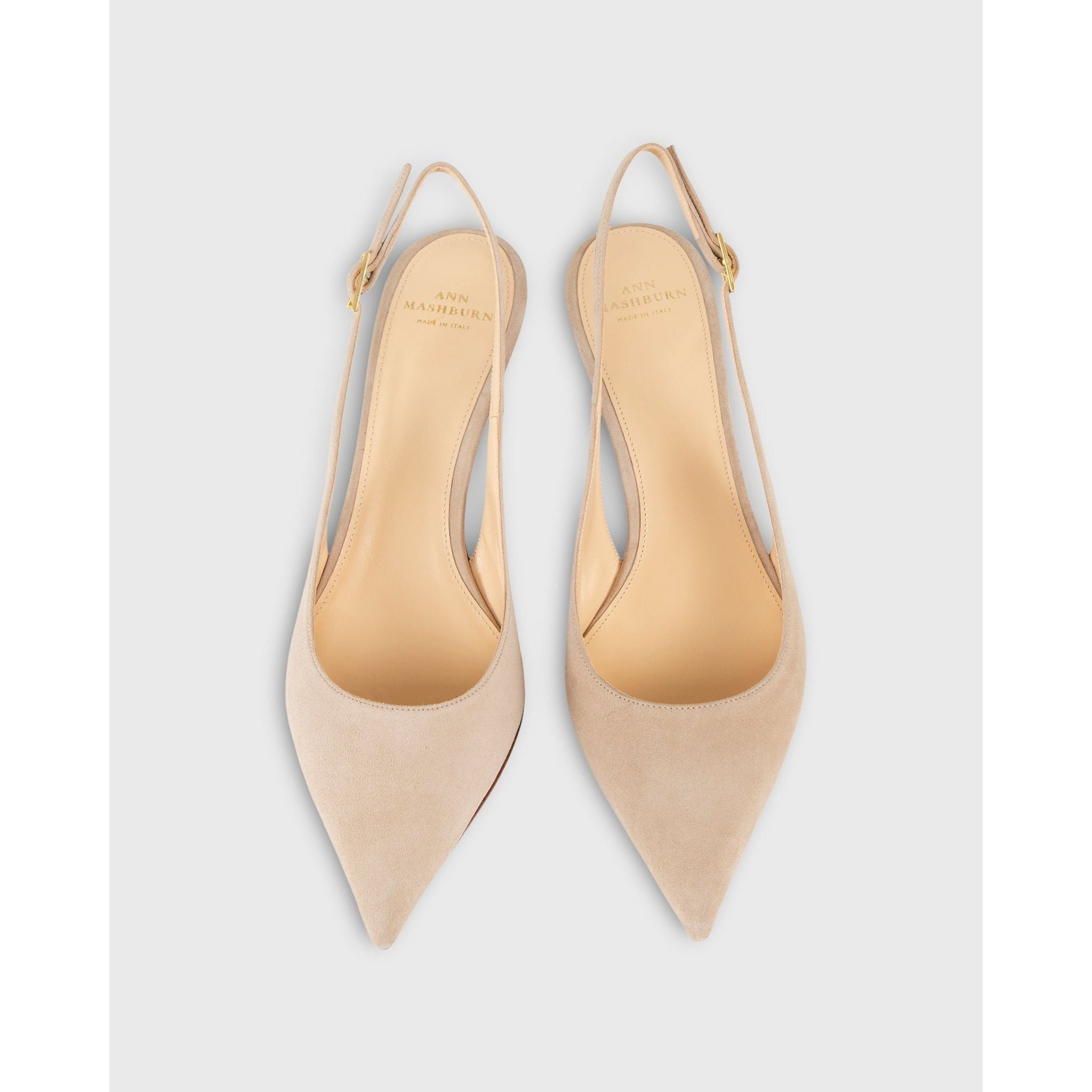 The Ann Mashburn Kitten Heel Slingback in pebble suede, featuring a pair of beige pointed-toe slingbacks highlighted by a small buckle on the straps, showcases retro charm against a plain white background. Crafted from lambskin leather, the insoles are adorned with "Ann Mashburn" printed in gold lettering.