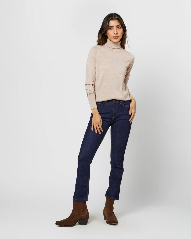 A person with long hair stands against a plain background wearing an Ann Mashburn Alida Turtleneck Sweater in heather oatmeal cashmere, dark blue jeans, and brown ankle boots. They are positioned slightly to the side and have their hands resting casually.