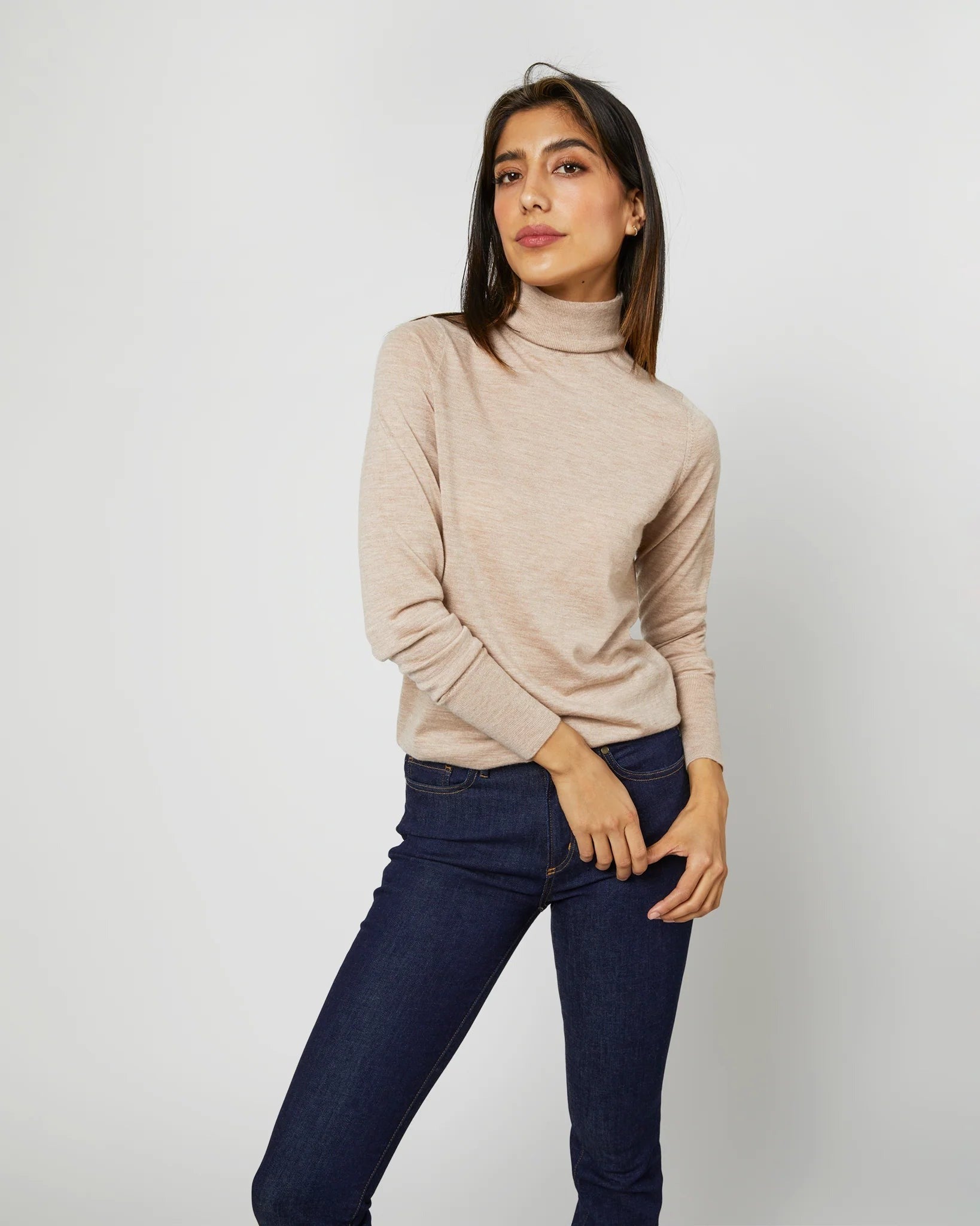 A woman with straight black hair stands against a plain background, wearing an Ann Mashburn Alida Turtleneck Sweater in Heather Oatmeal Cashmere and dark blue jeans. She has one hand resting on her hip and looks confidently at the camera, showcasing the elegance of versatile sweaters made from luxurious Mongolian cashmere.