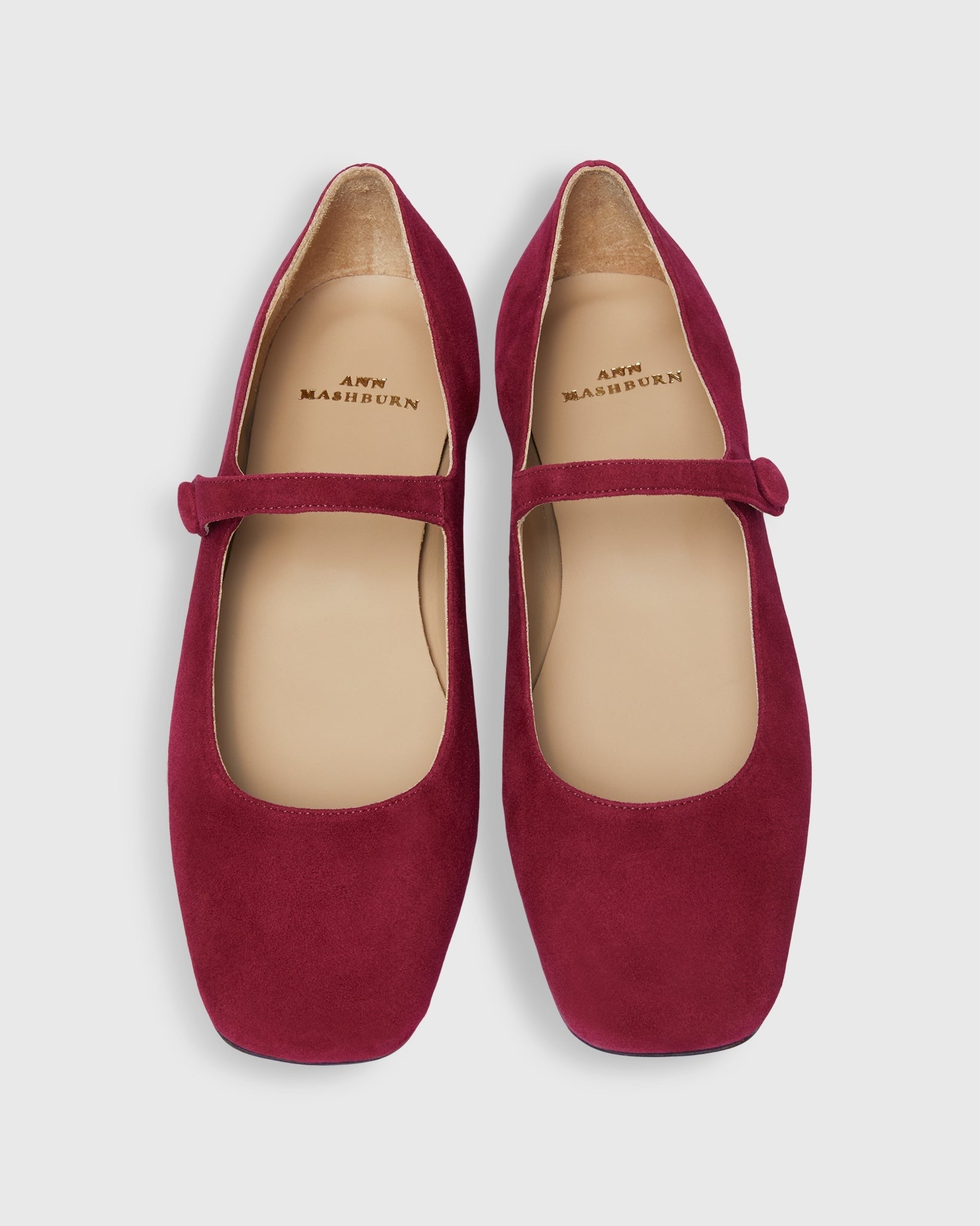 A pair of Ann Mashburn Square Toe Mary Jane flats in rich beet suede, crafted from luxurious Italian calf suede leather, featuring beige insoles and a single strap across each shoe. The brand name "Ann Mashburn" is elegantly printed on the insoles. These stylish shoes are displayed against a pristine white background.