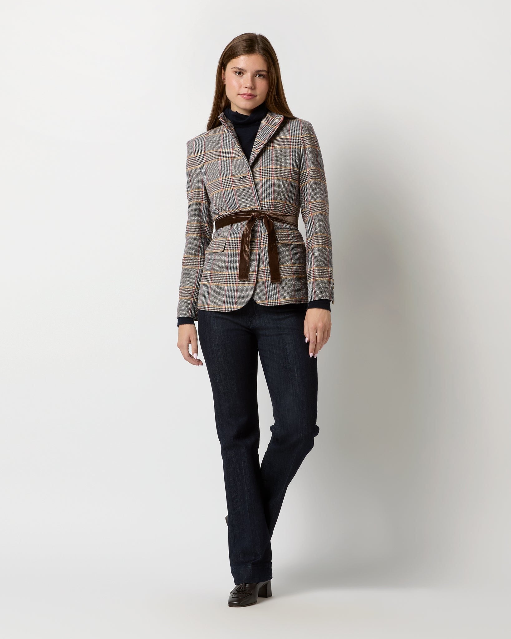 A woman stands confidently, wearing an Ann Mashburn Amelia Blazer in Glen Plaid, cinched at the waist with a brown belt. She pairs it with a black turtleneck, dark jeans, and black shoes. Her straight hair frames her face as she gives a slight smile against a plain light gray background.