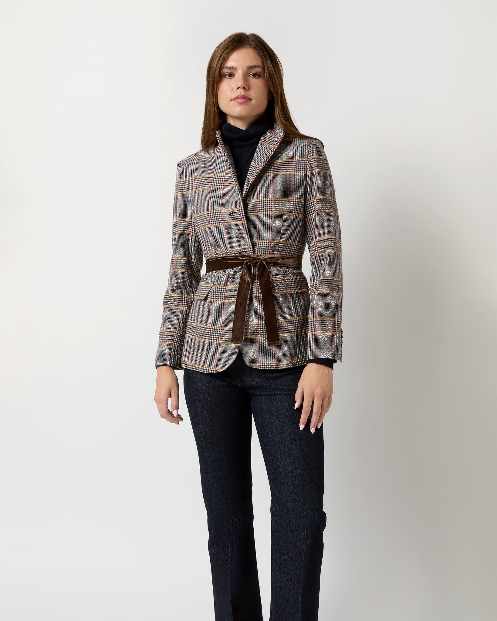 A person wearing the Ann Mashburn Amelia Blazer Glen Plaid, crafted from an Italian wool blend and belted at the waist with a velvet ribbon belt over a black turtleneck sweater and dark trousers, stands against a plain, light background. Their expression is neutral, and their arms are relaxed by their sides.