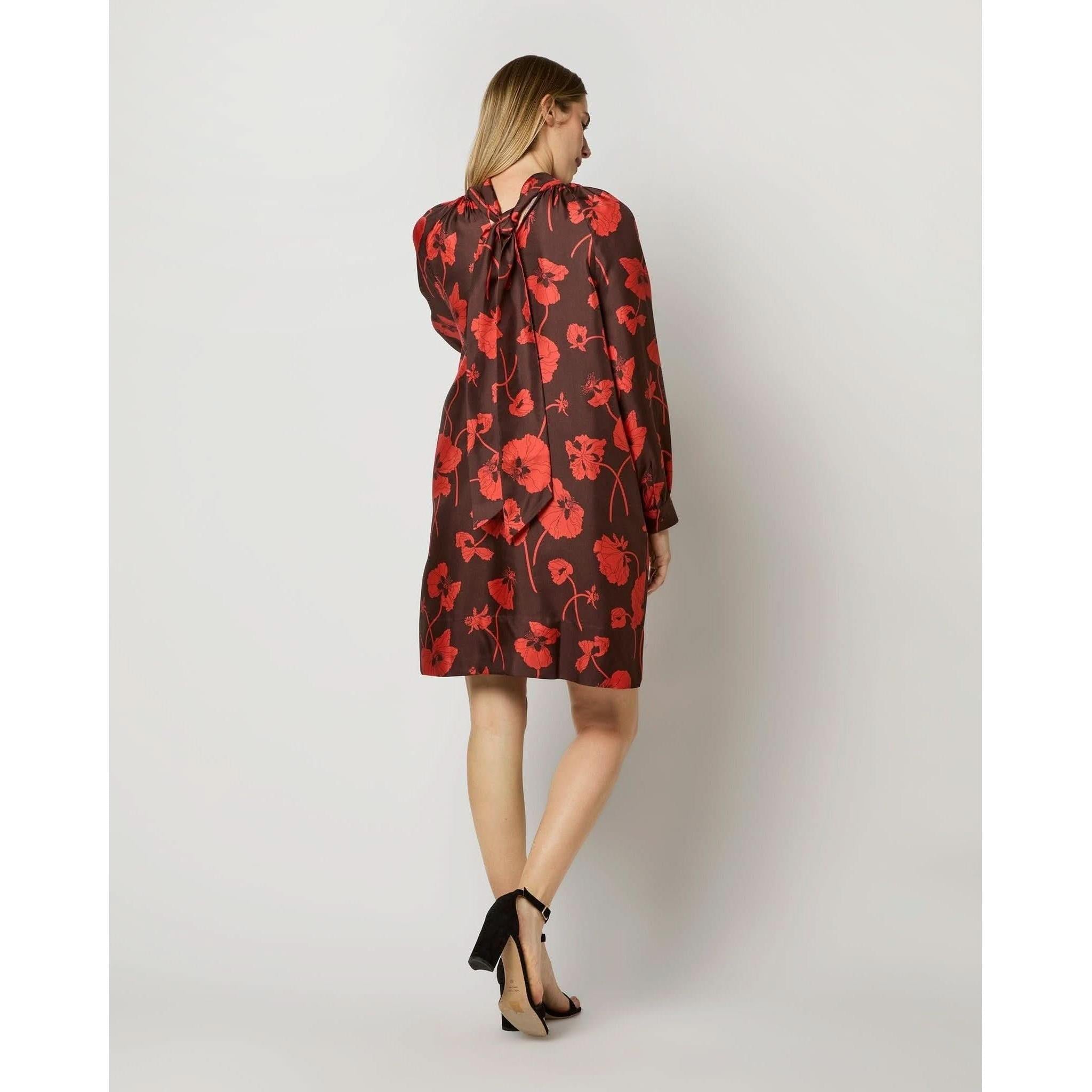 A woman stands facing away, showcasing the Ann Mashburn Maggie Dress. This elegant autumn dress by Ann Mashburn features long sleeves and a red floral pattern on dark brown silk fabric from Liberty Fabrics. The dress includes a stylish bow tie at the back of the neck. She is wearing black high-heeled shoes, and her long, light brown hair cascades down against a plain, light-colored background.