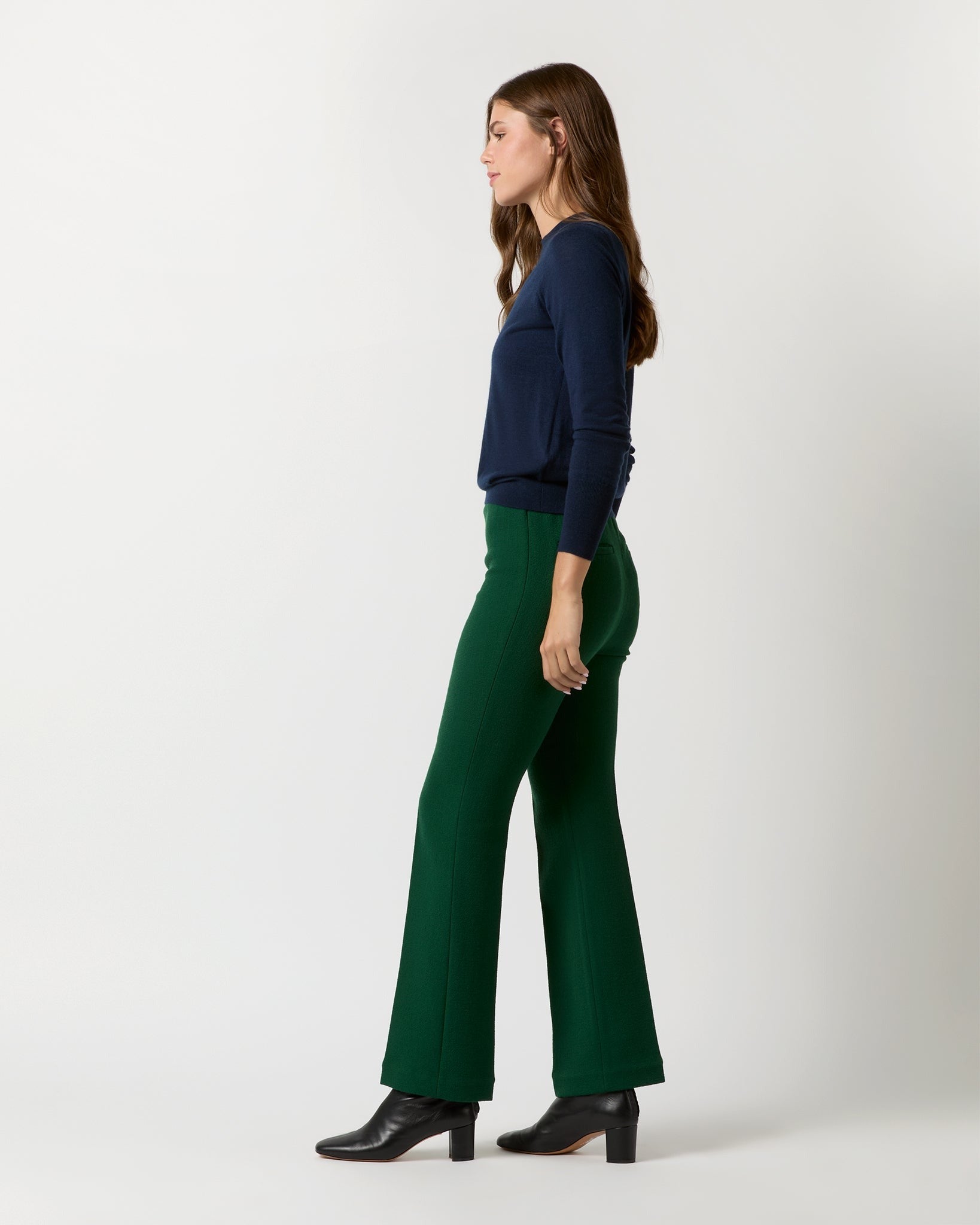 A woman standing in profile against a plain light background, wearing a dark blue long-sleeve top, Ann Mashburn's Alice Pant in Forest Double Face Crepe, and black heeled ankle boots. She has long brown hair and is looking forward with her arms relaxed by her sides.