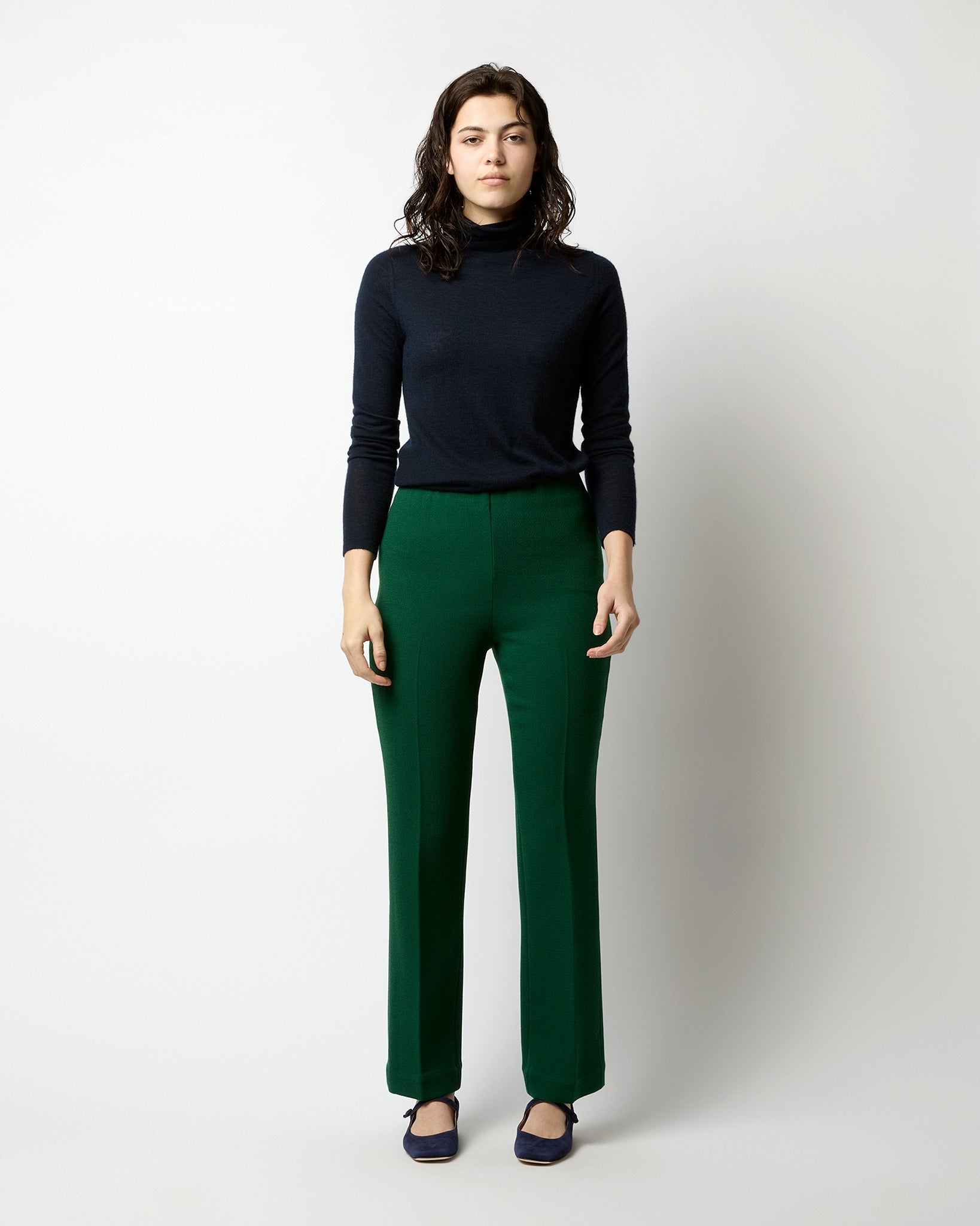 A person with medium-length, wavy dark hair is standing against a plain background. They are wearing a black turtleneck sweater, the Ann Mashburn Alice Pant in Forest Double Face Crepe with an invisible center back zipper, and dark shoes. Their arms are relaxed by their sides.