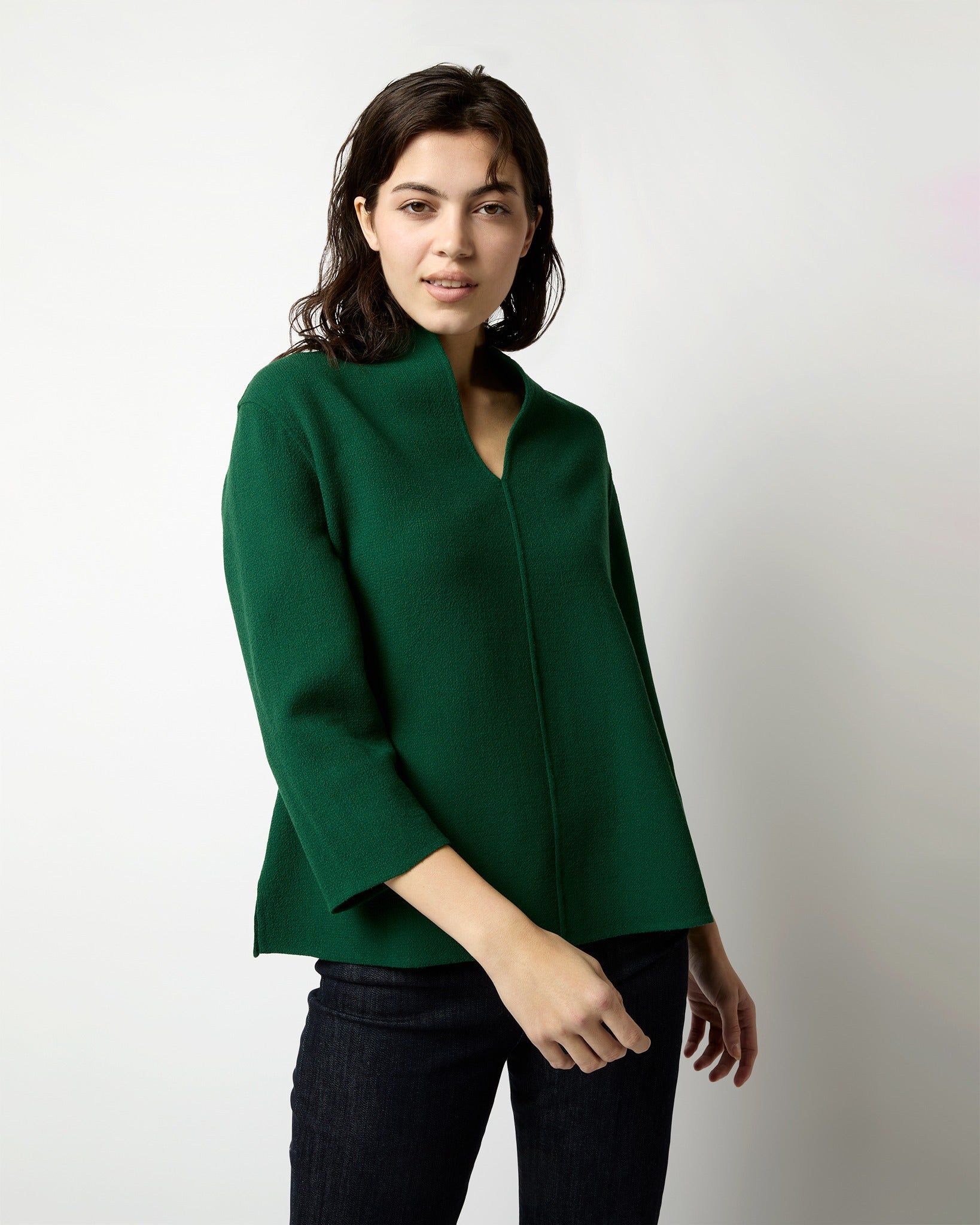 A person with shoulder-length dark hair is wearing the Ann Mashburn Camilla Top in Forest Double Face Stretch Crepe, which features long sleeves and a V-neck design. They are standing against a plain white background, looking directly at the camera with a neutral expression.