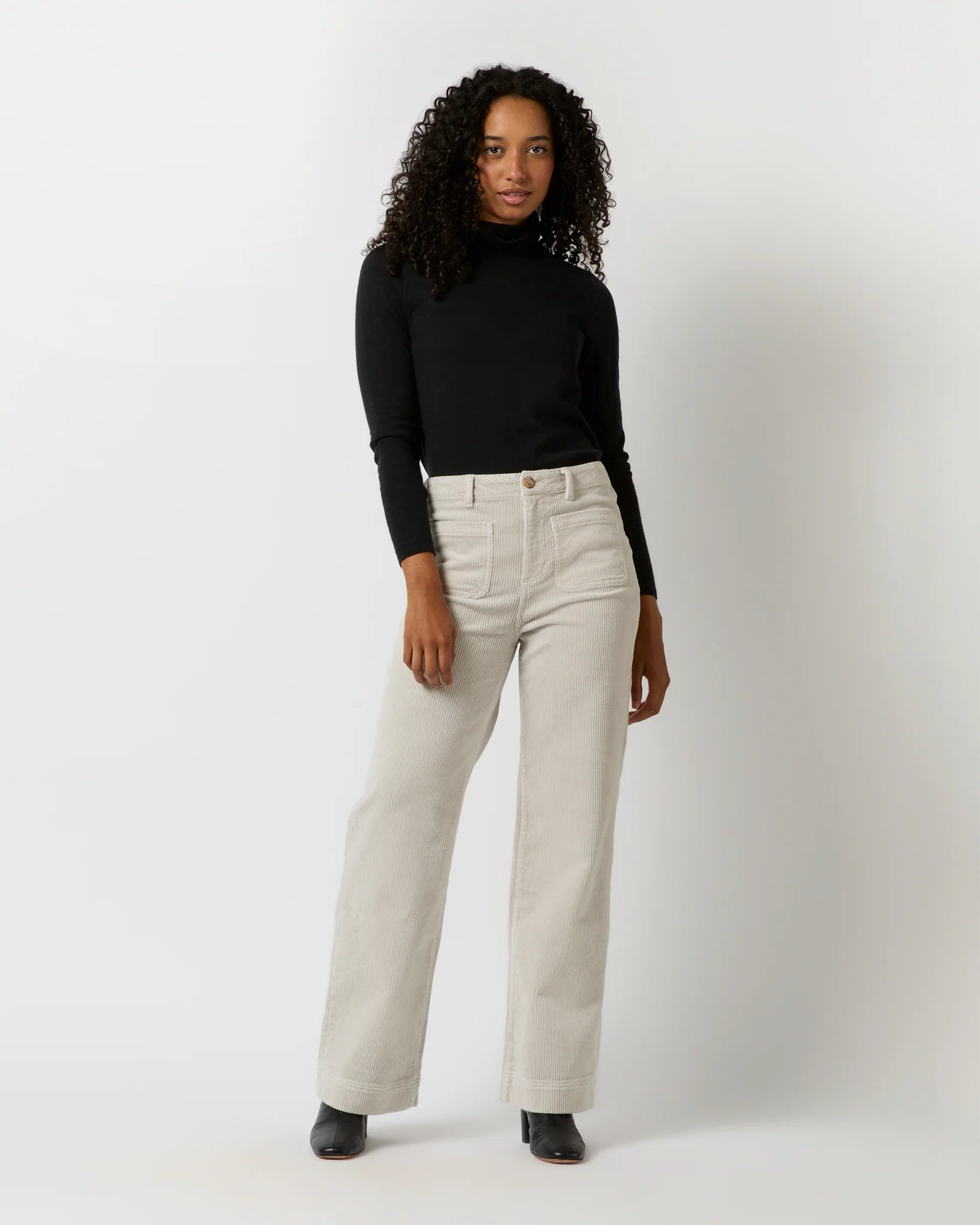 A person with curly hair is standing against a light background, facing away from the camera. They are wearing a black long-sleeve top, Ann Mashburn Coco Patch Pocket Jean in a cream-colored wide-leg design that offers a flattering fit, and black shoes. Their hands are resting on their hips.