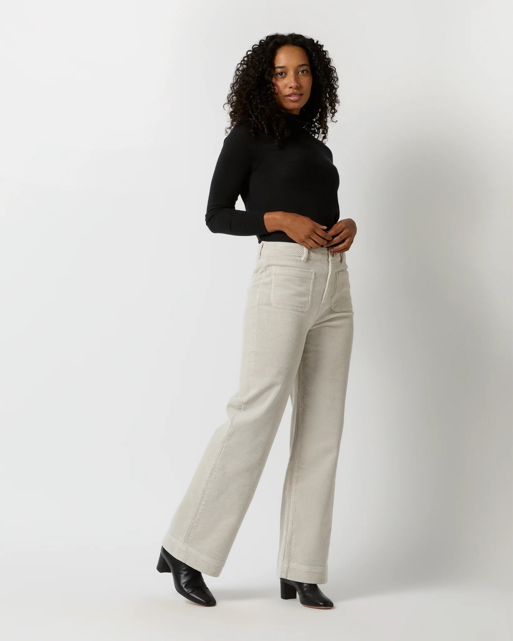 A person with curly hair is standing against a light background, facing away from the camera. They are wearing a black long-sleeve top, Ann Mashburn Coco Patch Pocket Jean in a cream-colored wide-leg design that offers a flattering fit, and black shoes. Their hands are resting on their hips.