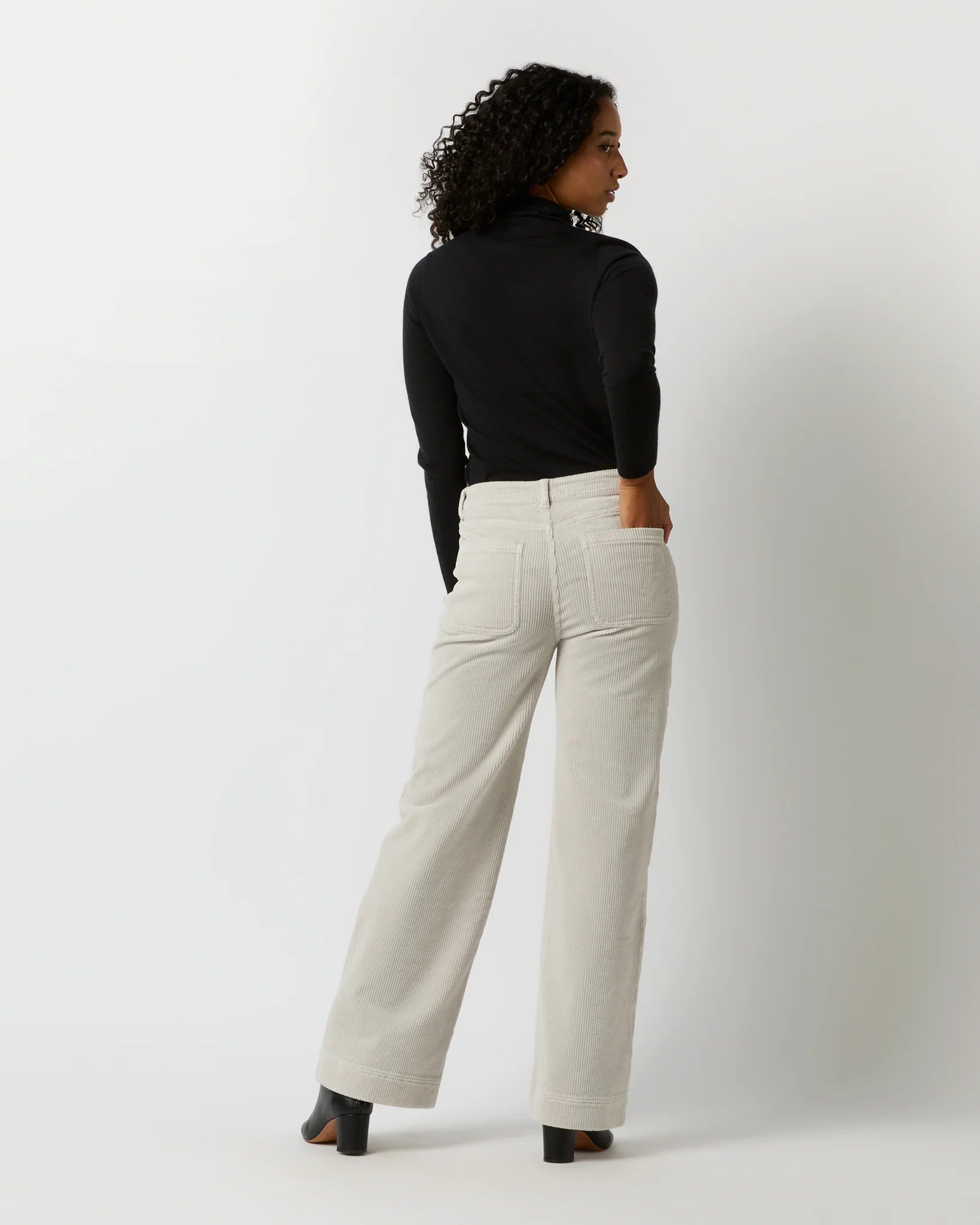 A person with curly hair is standing against a light background, facing away from the camera. They are wearing a black long-sleeve top, Ann Mashburn Coco Patch Pocket Jean in a cream-colored wide-leg design that offers a flattering fit, and black shoes. Their hands are resting on their hips.