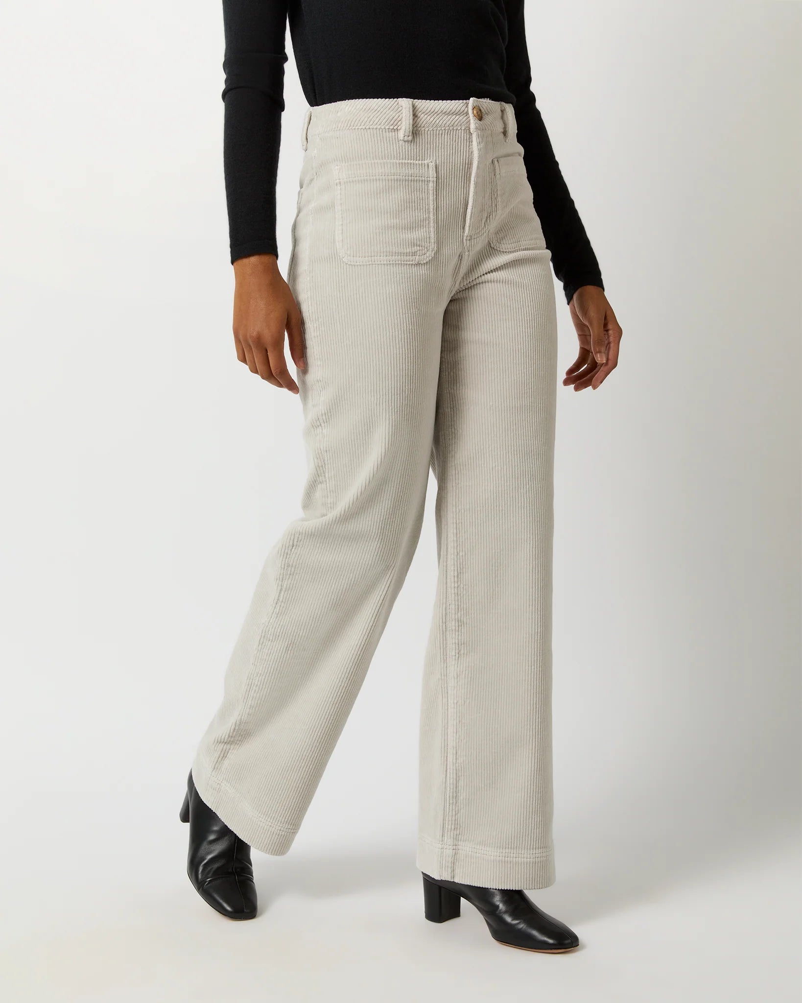 A person is wearing Ann Mashburn Coco Patch Pocket Jeans in light beige corduroy with wide legs, paired with a black long-sleeve top and black heeled boots. The flattering fit of the outfit accentuates their style, and only the lower half of the body is visible against a plain white background.