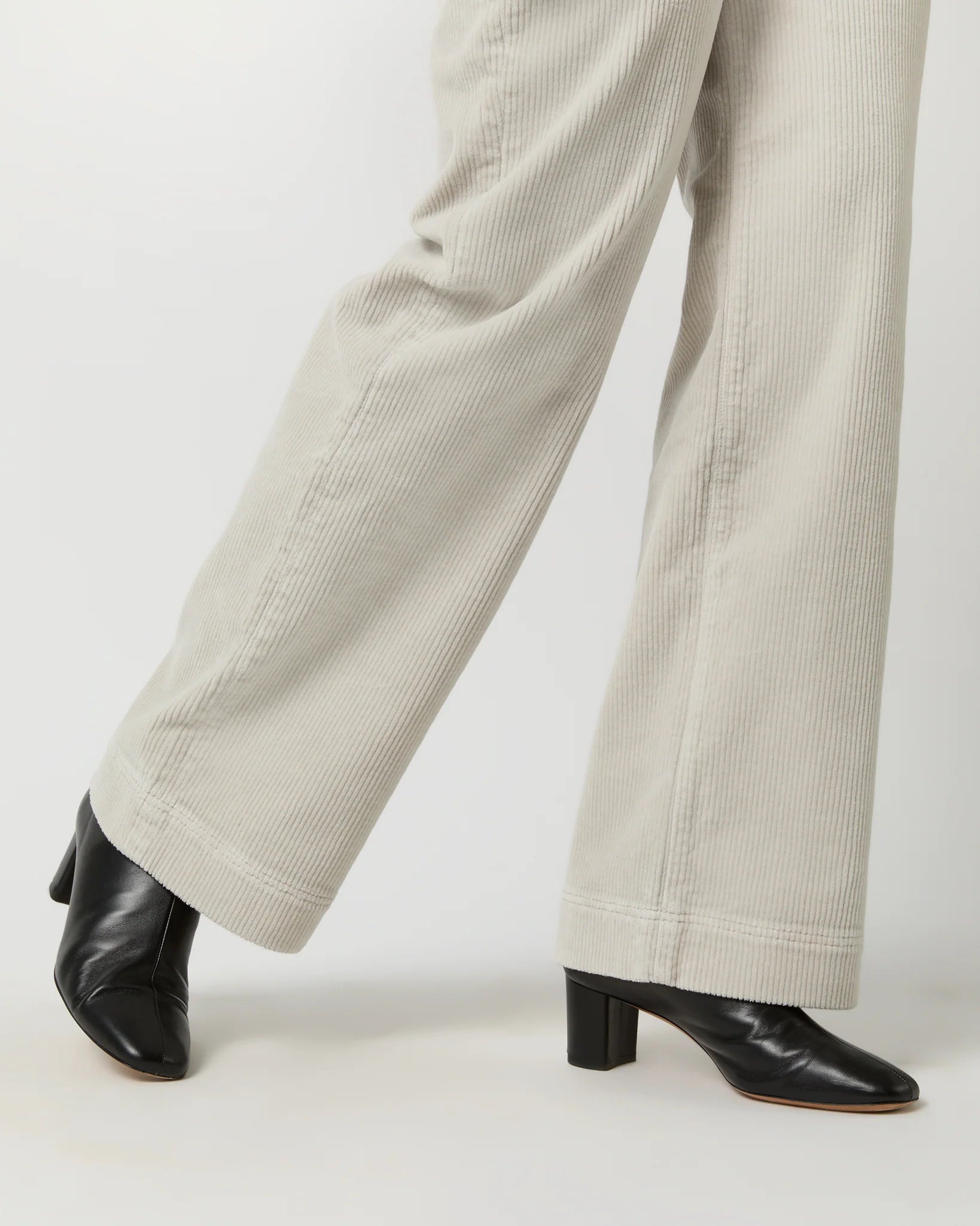 A person with curly hair is standing against a light background, facing away from the camera. They are wearing a black long-sleeve top, Ann Mashburn Coco Patch Pocket Jean in a cream-colored wide-leg design that offers a flattering fit, and black shoes. Their hands are resting on their hips.