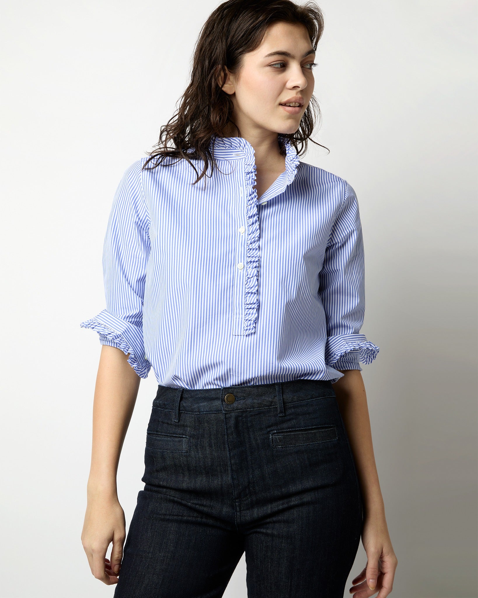 A person with wavy dark hair is wearing the Ann Mashburn Frill Shirt in Blue Bengal Stripe Poplin, featuring ruffled detailing on the front and sleeves. Paired with dark jeans, they are standing against a plain white background, looking slightly to the side and smiling.