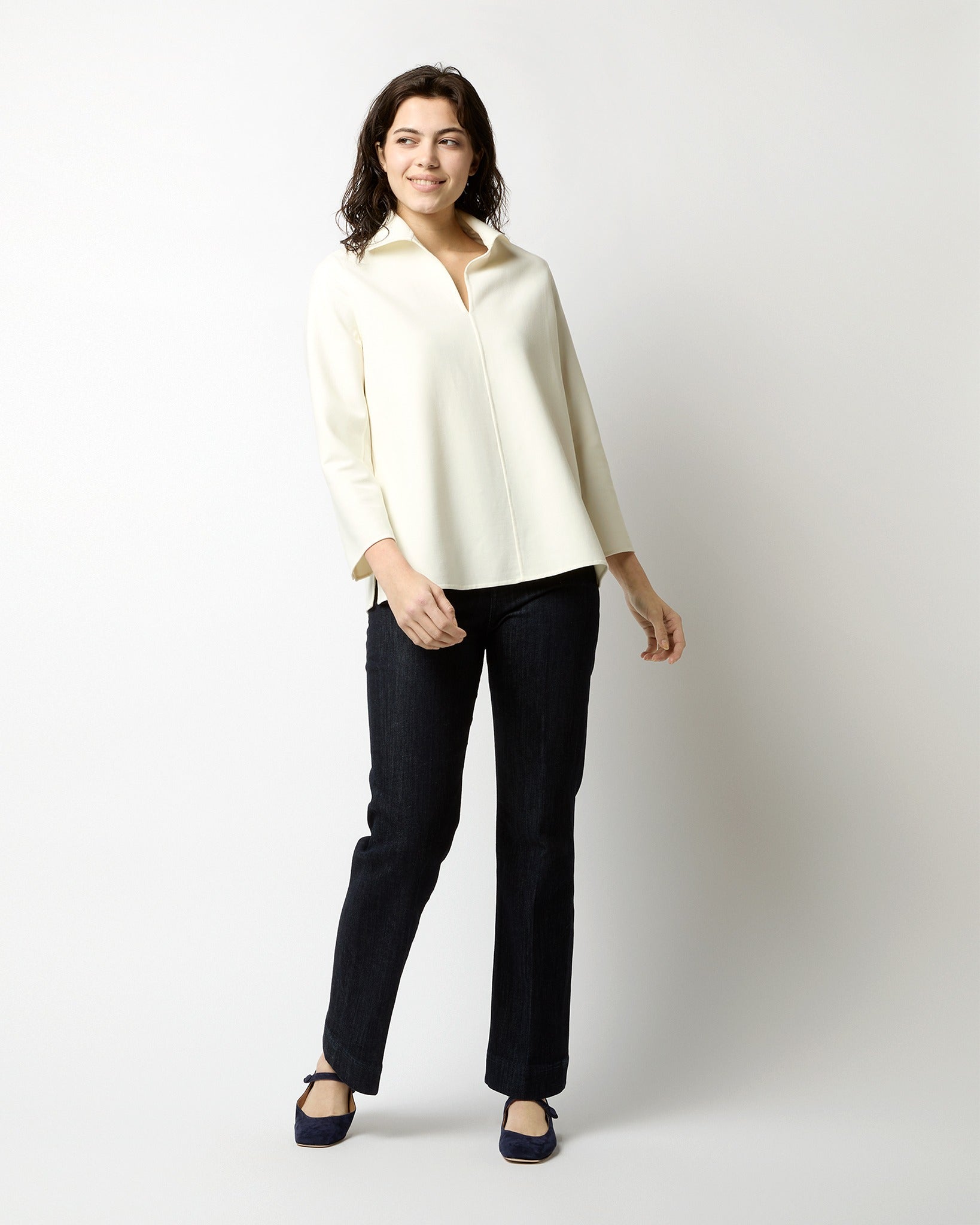 A person with medium-length, dark hair stands against a plain white background. They are wearing an Ann Mashburn Olympia Top in Ivory Bi-Stretch Double Face with a wide collar and dark straight-leg pants. Smiling slightly, they pose with one hand hanging by their side and the other extended out.