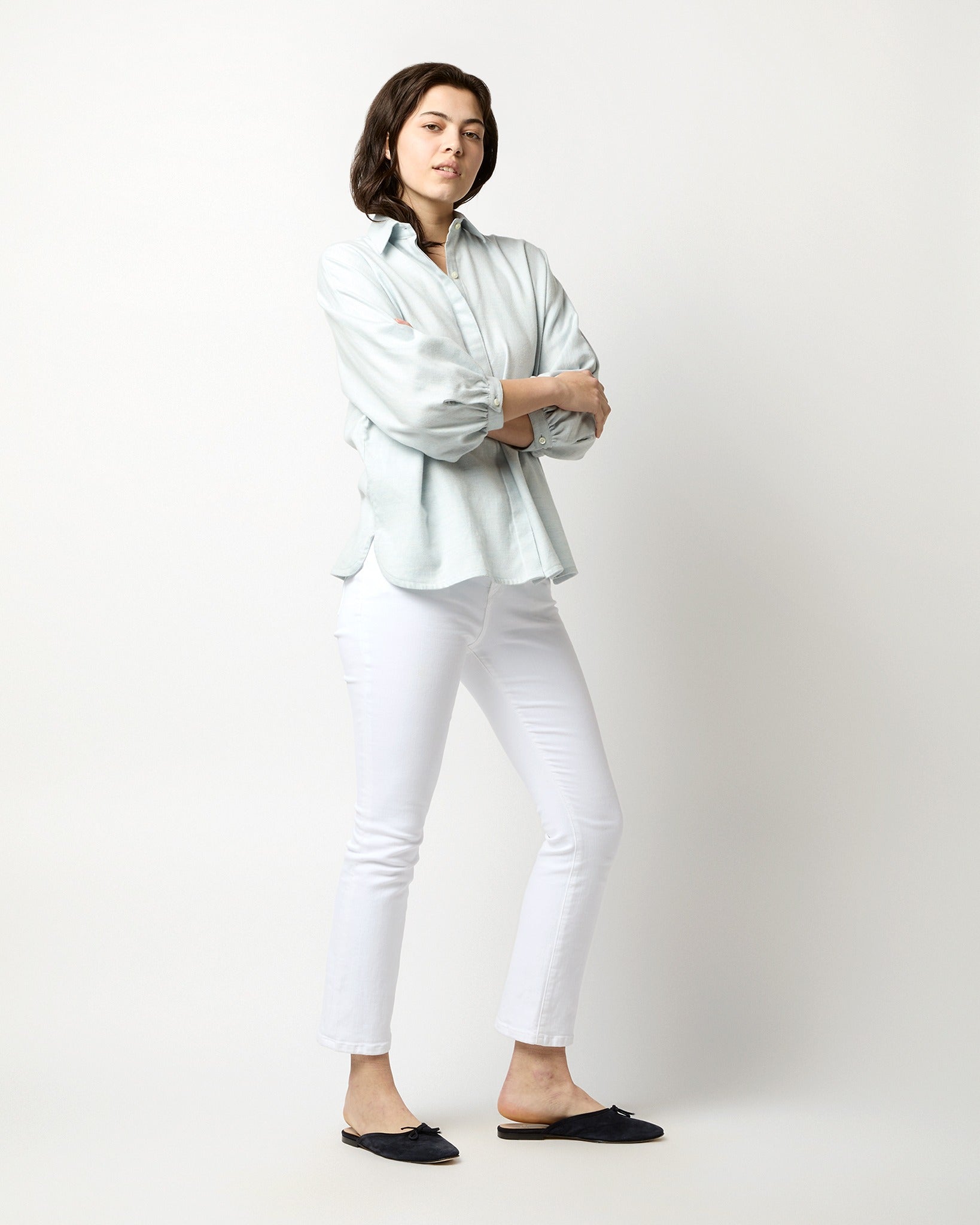 A person with short dark hair stands against a plain white background, wearing an Ann Mashburn Volume Kimono Shirt in Sky Brushed Herringbone, white skinny jeans, and black slip-on shoes. They have their arms crossed and are slightly leaning to one side.