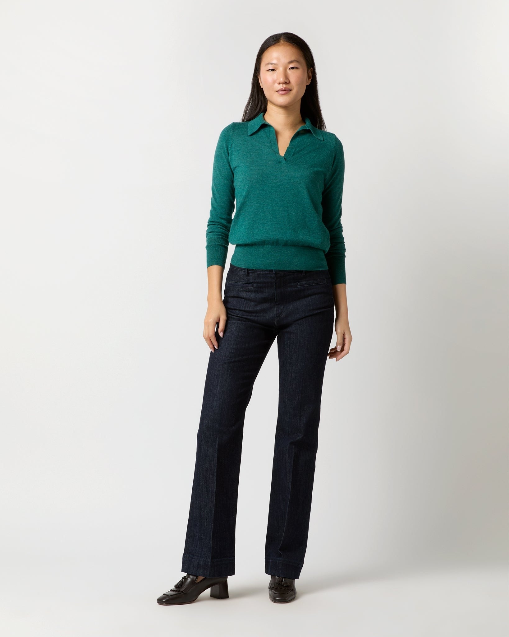 A person stands against a plain white background, wearing an Ann Mashburn Alida Johnny-Collar Sweater in Highland Green Cashmere with a v-neck and dark blue jeans. They have long dark hair and are wearing black heeled shoes. They are facing forward with a neutral expression and their arms relaxed by their sides.