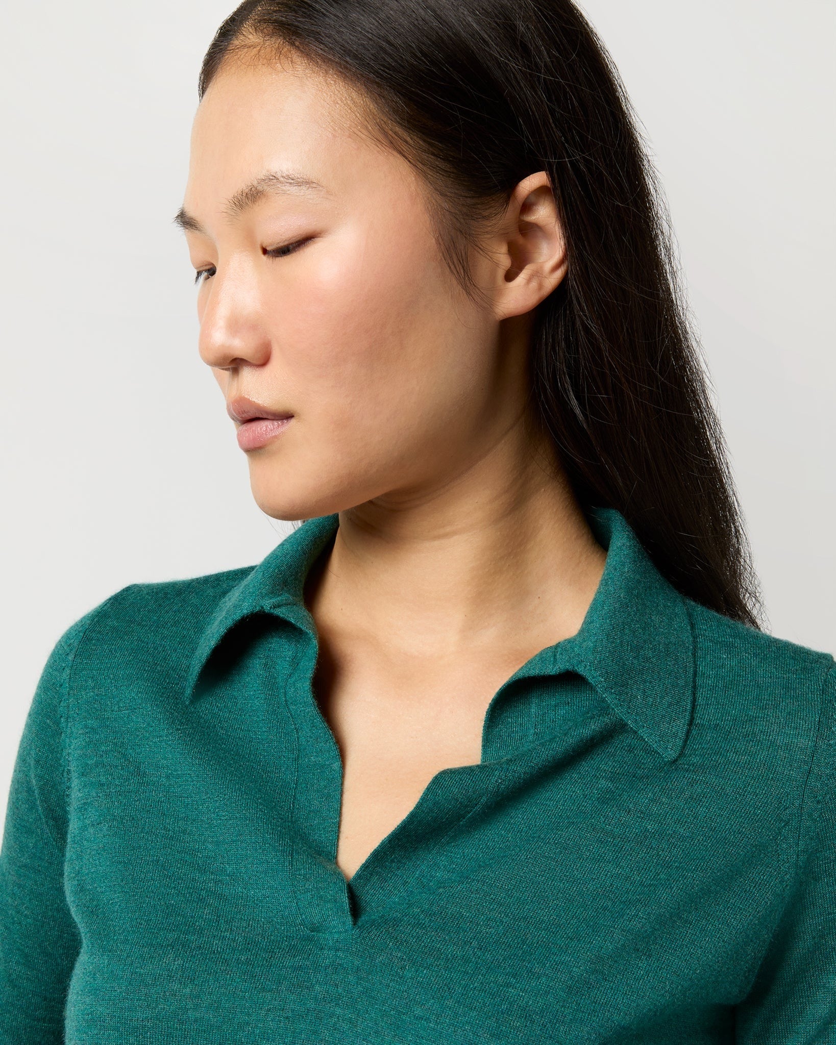 A person with long dark hair is shown in a mid-shot looking downward to their right. They are wearing an Ann Mashburn Alida Johnny-Collar Sweater in Highland Green Cashmere and have a calm expression on their face. The background is a plain, light color.