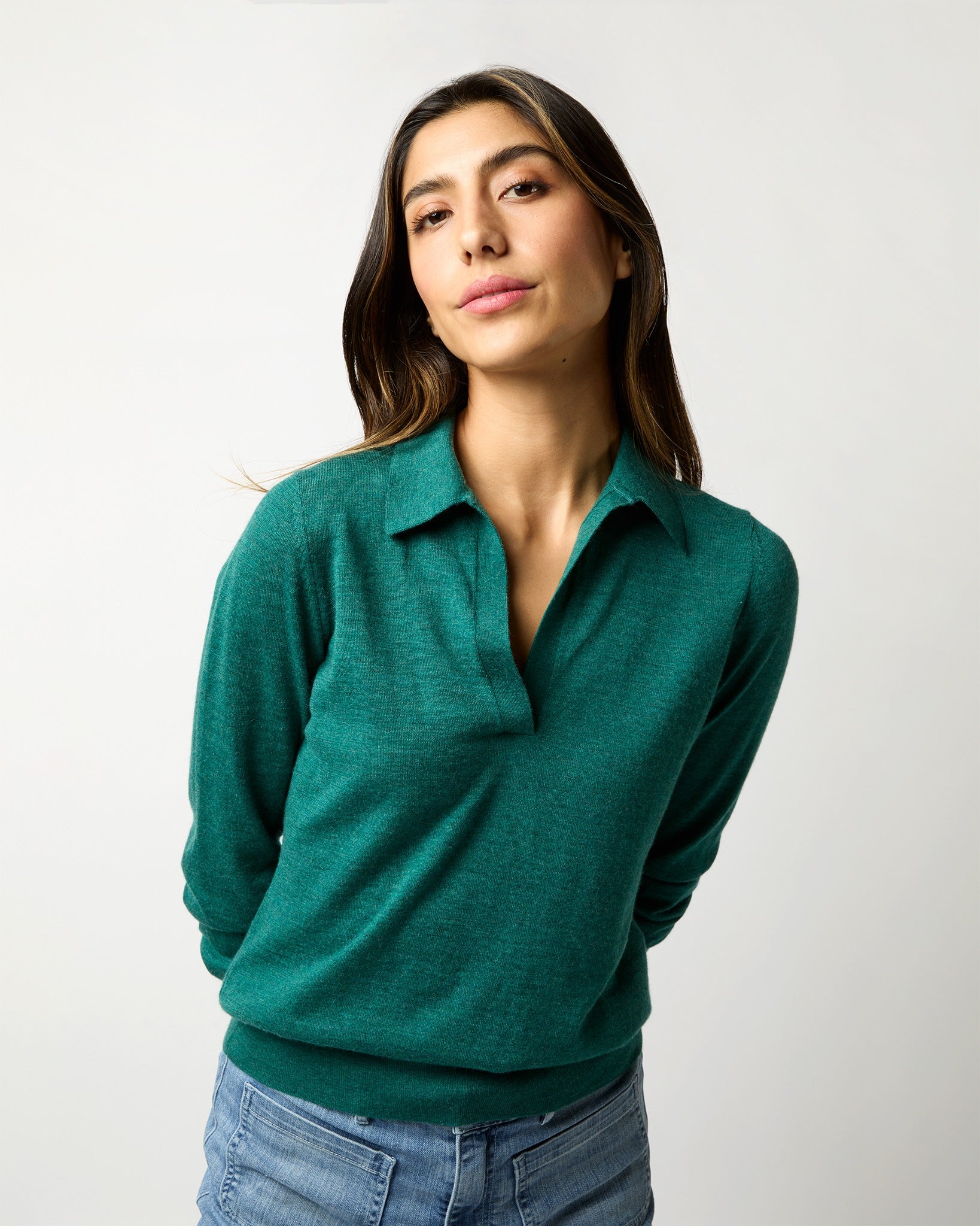 A person with long dark hair, wearing the Ann Mashburn Alida Johnny-Collar Sweater in Highland Green Cashmere and blue jeans, poses against a plain, light grey background. They have a relaxed expression and have their hands behind their back.