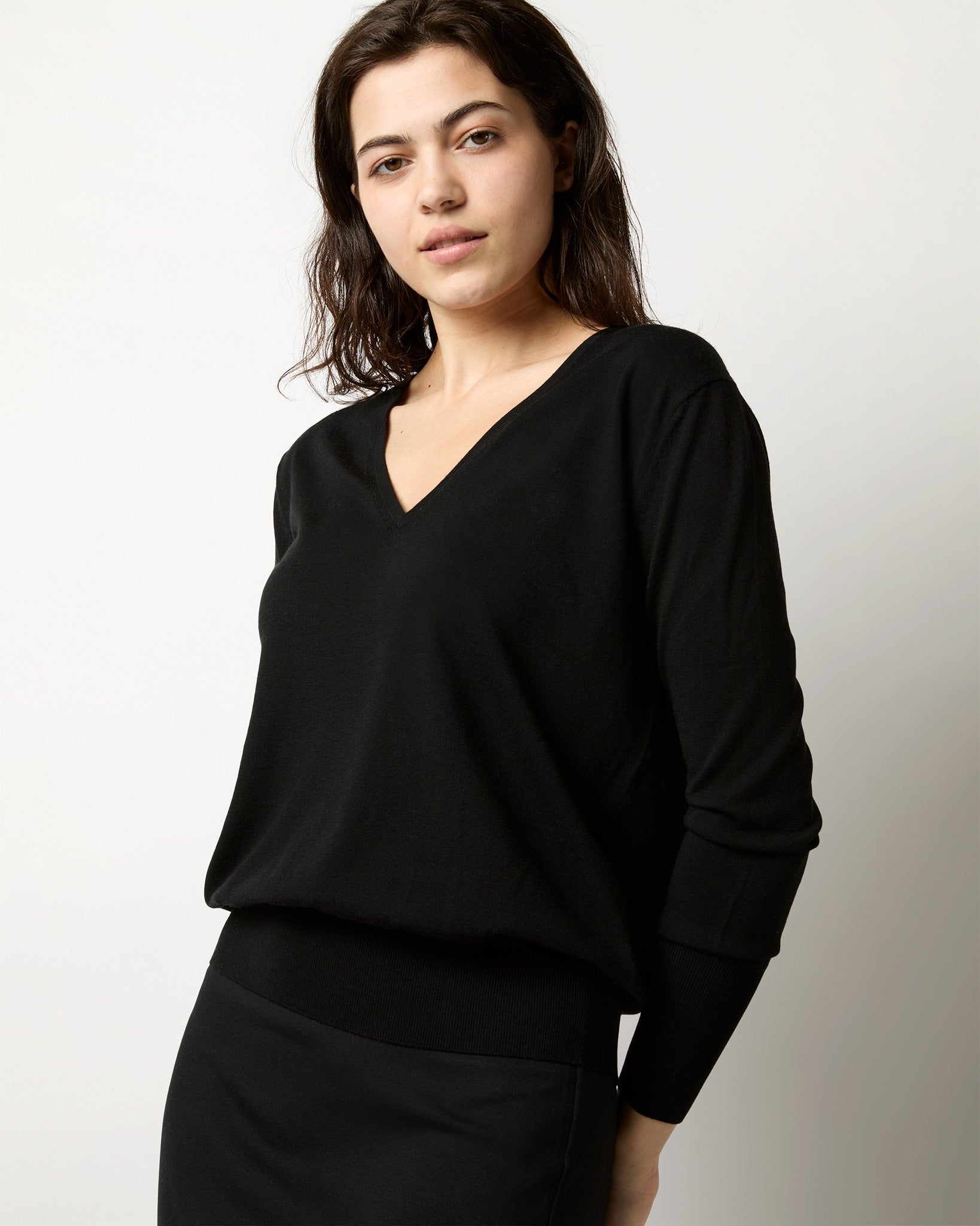 A woman with wavy dark hair wears an Ann Mashburn Kate V-Neck Sweater in Black Escorial Wool and a matching black skirt, posing against a plain light background.