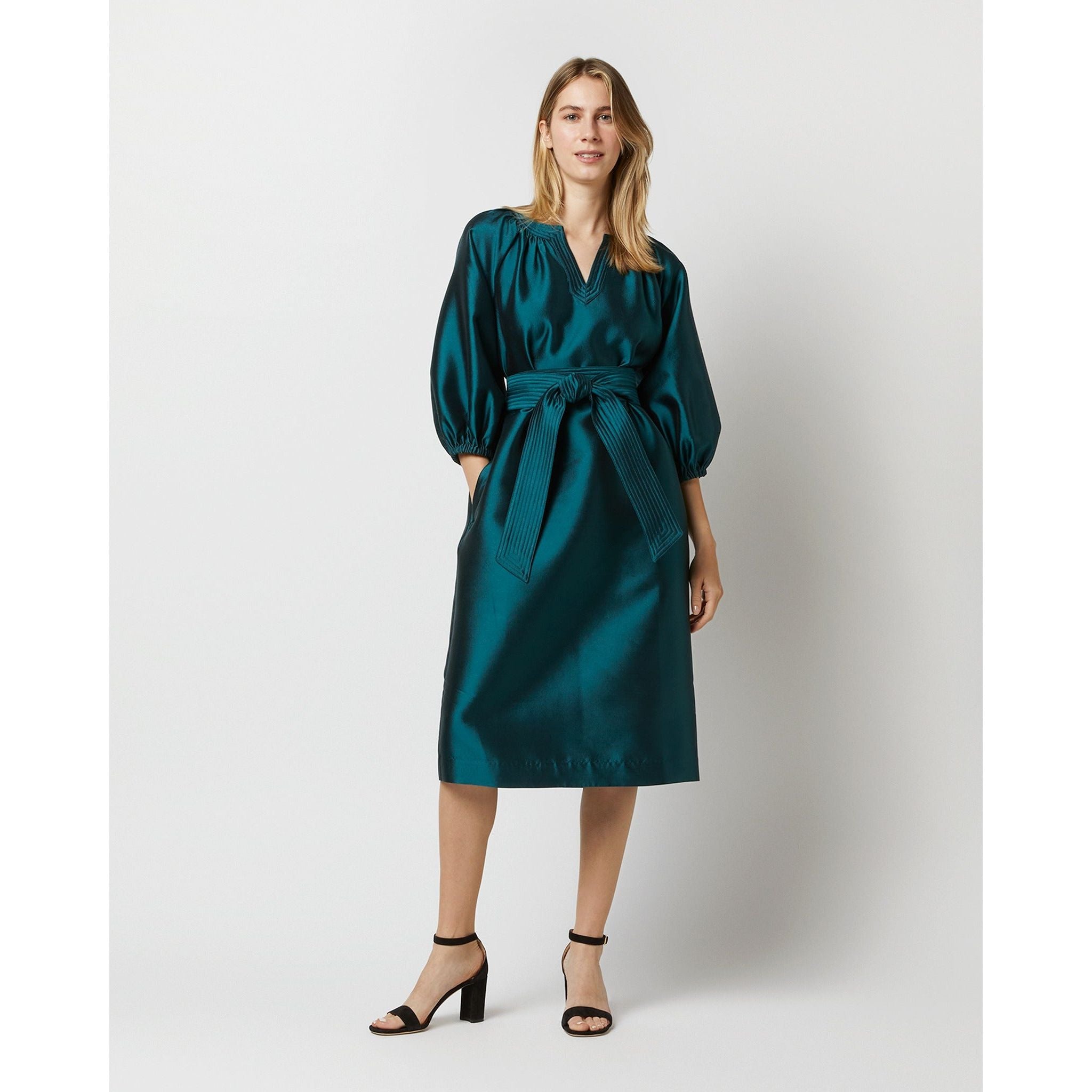 A person is wearing the Ann Mashburn Trapunto Blouson Dress in peacock silk wool, featuring 3/4 blouson sleeves and a matching belt. With long hair, they stand with one hand on their hip, styled in black open-toe block heels against a plain white background.