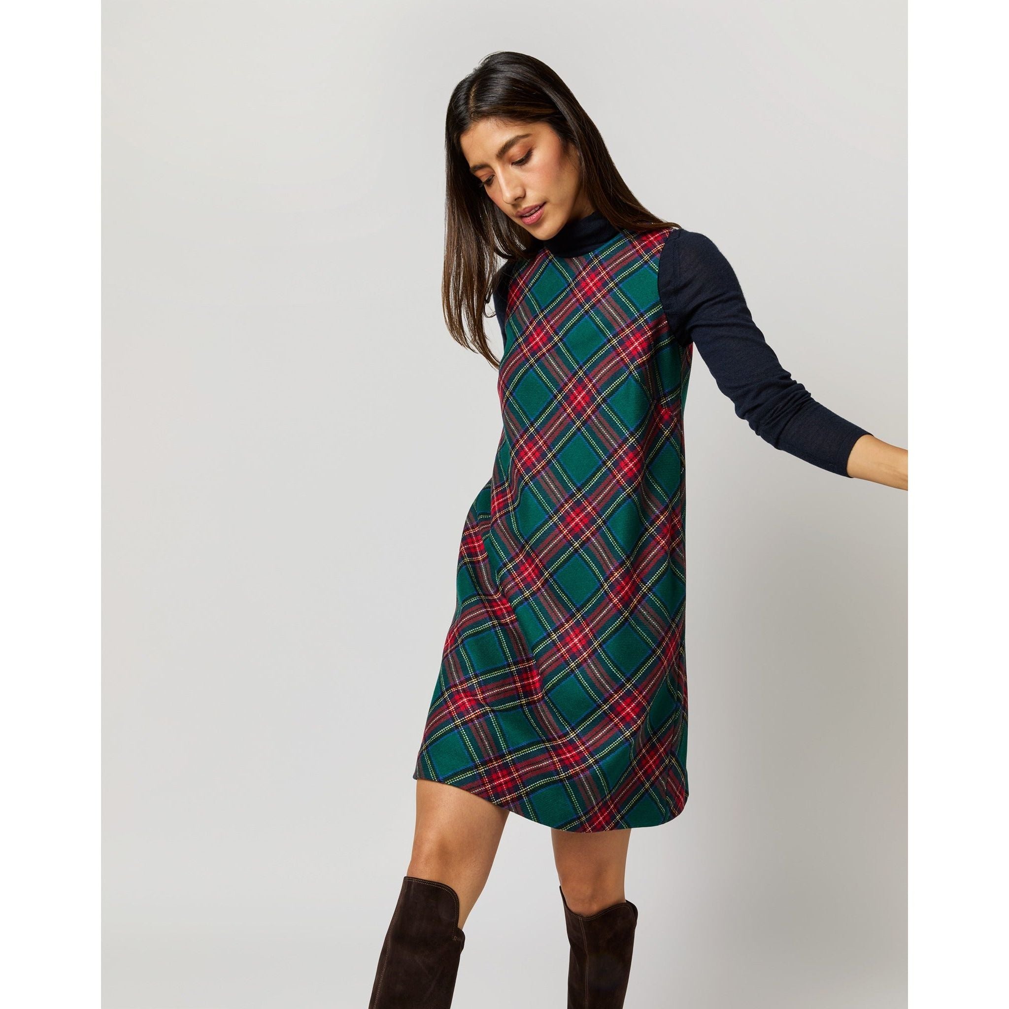 A woman models the Ann Mashburn Susy Shift Dress in Green/Red Royal Stewart Tartan, crafted from soft wool and worn over a dark long-sleeve shirt. Her head is slightly tilted, and she pairs the outfit with knee-high black boots against a plain white background.