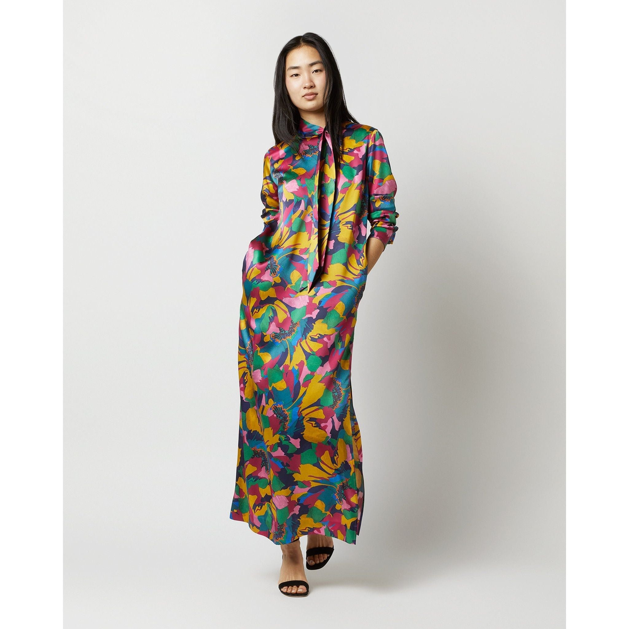 A person with long hair is wearing the Ann Mashburn Leigh Dress, which features a vibrant floral pattern on silk shantung. They have their hands tucked into the dress pockets while standing against a plain white background, and are paired with black heeled sandals.