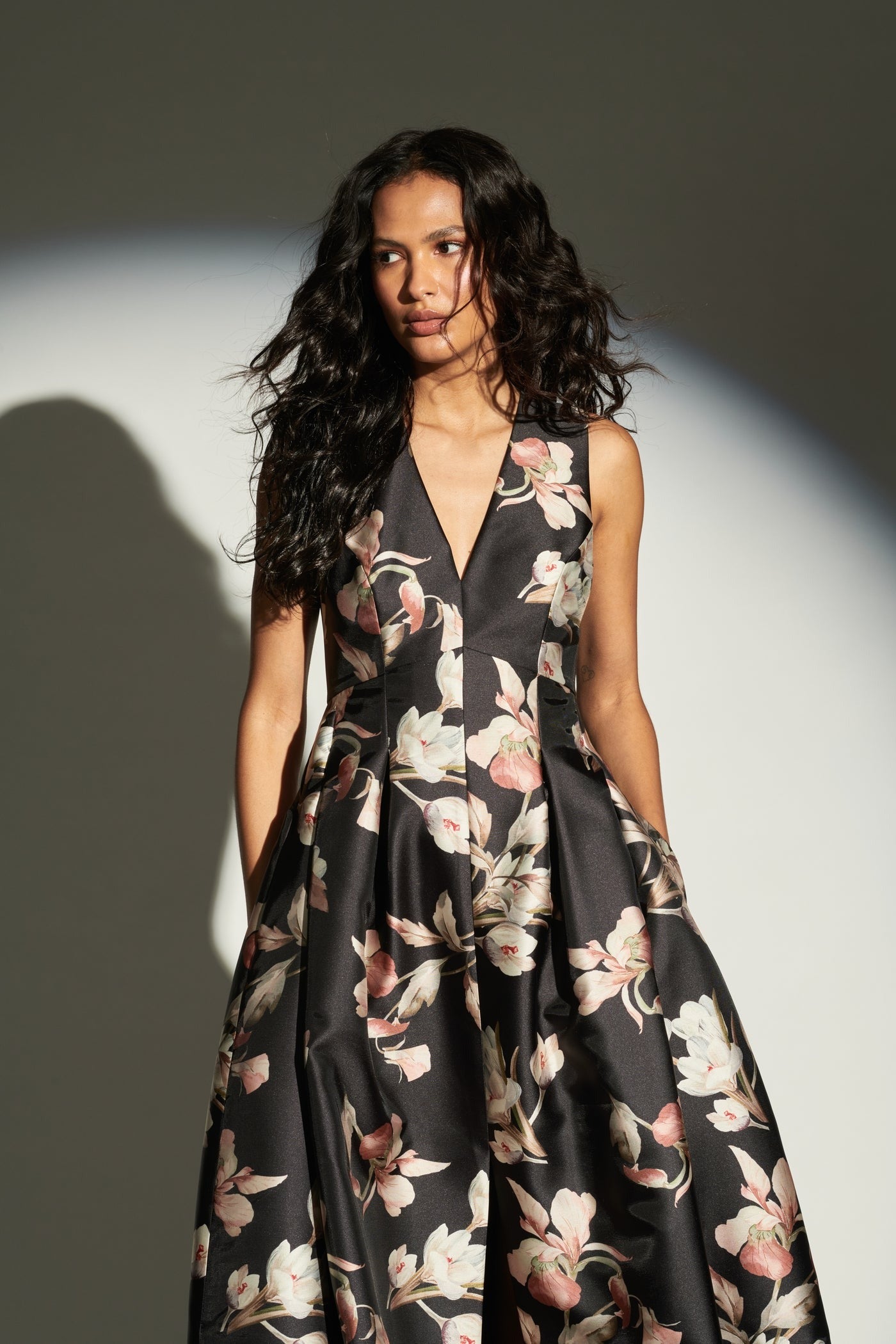 A woman with long, wavy hair and a classic silhouette is wearing the Sachin & Babi Brooke Gown - Tonal Petal, a sleeveless black dress adorned with a white and pink floral pattern. She stands against a neutral background, her shadow cast behind her.