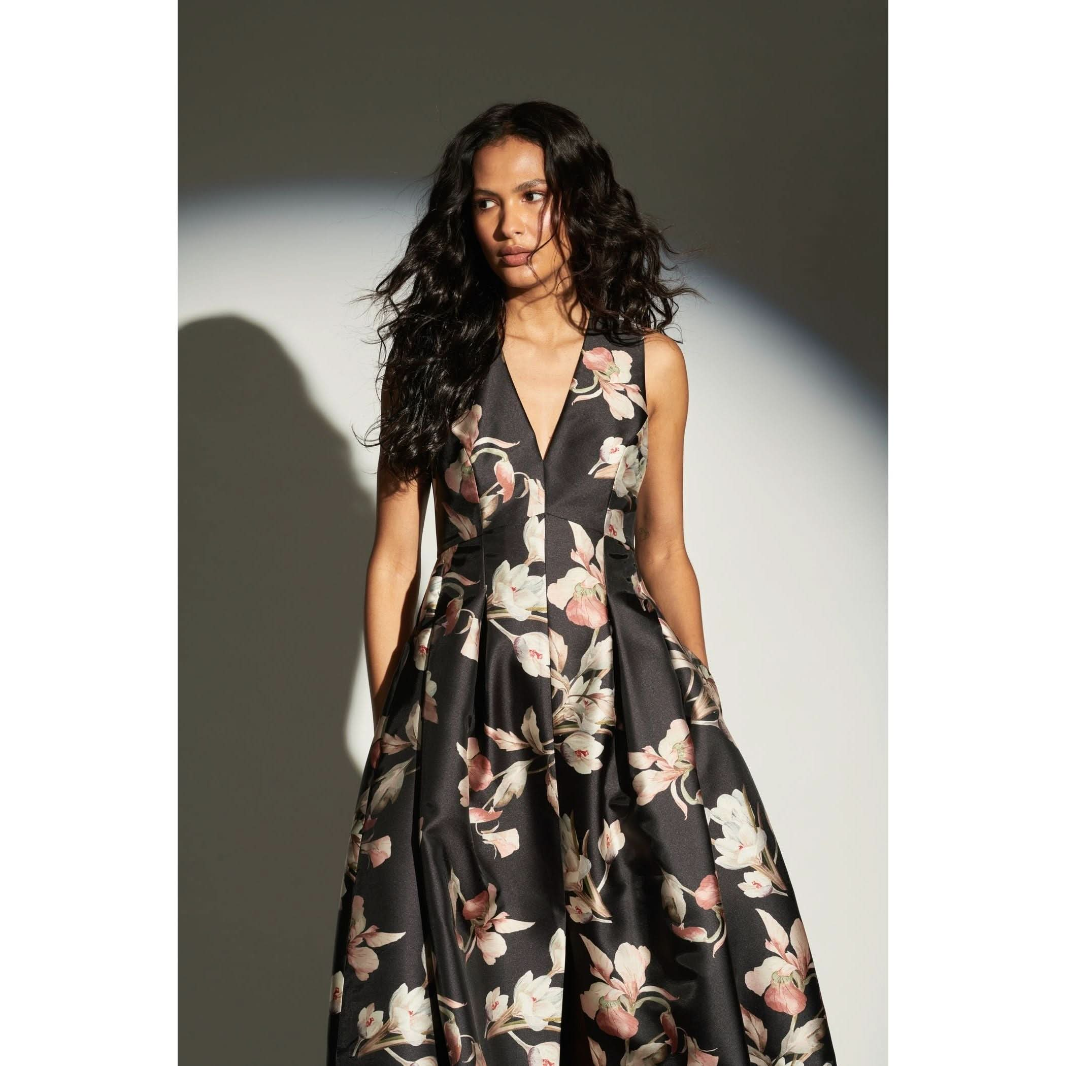 A woman with long, wavy hair and a classic silhouette is wearing the Sachin & Babi Brooke Gown - Tonal Petal, a sleeveless black dress adorned with a white and pink floral pattern. She stands against a neutral background, her shadow cast behind her.