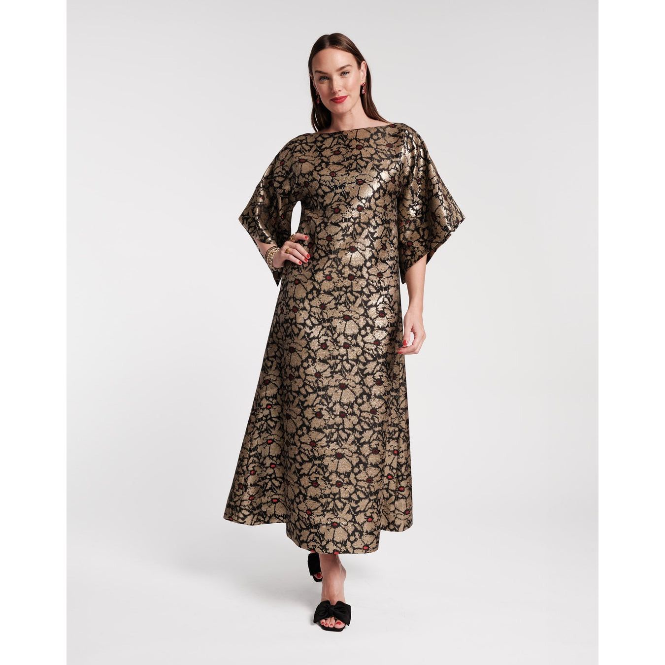 A woman wearing the Frances Valentine Spinnaker Maxi Dress in Winter Anemone Jacquard Black/Gold poses against a white background. The dress showcases a metallic floral pattern and bell sleeves. She complements her look with black shoes and has long, dark hair.