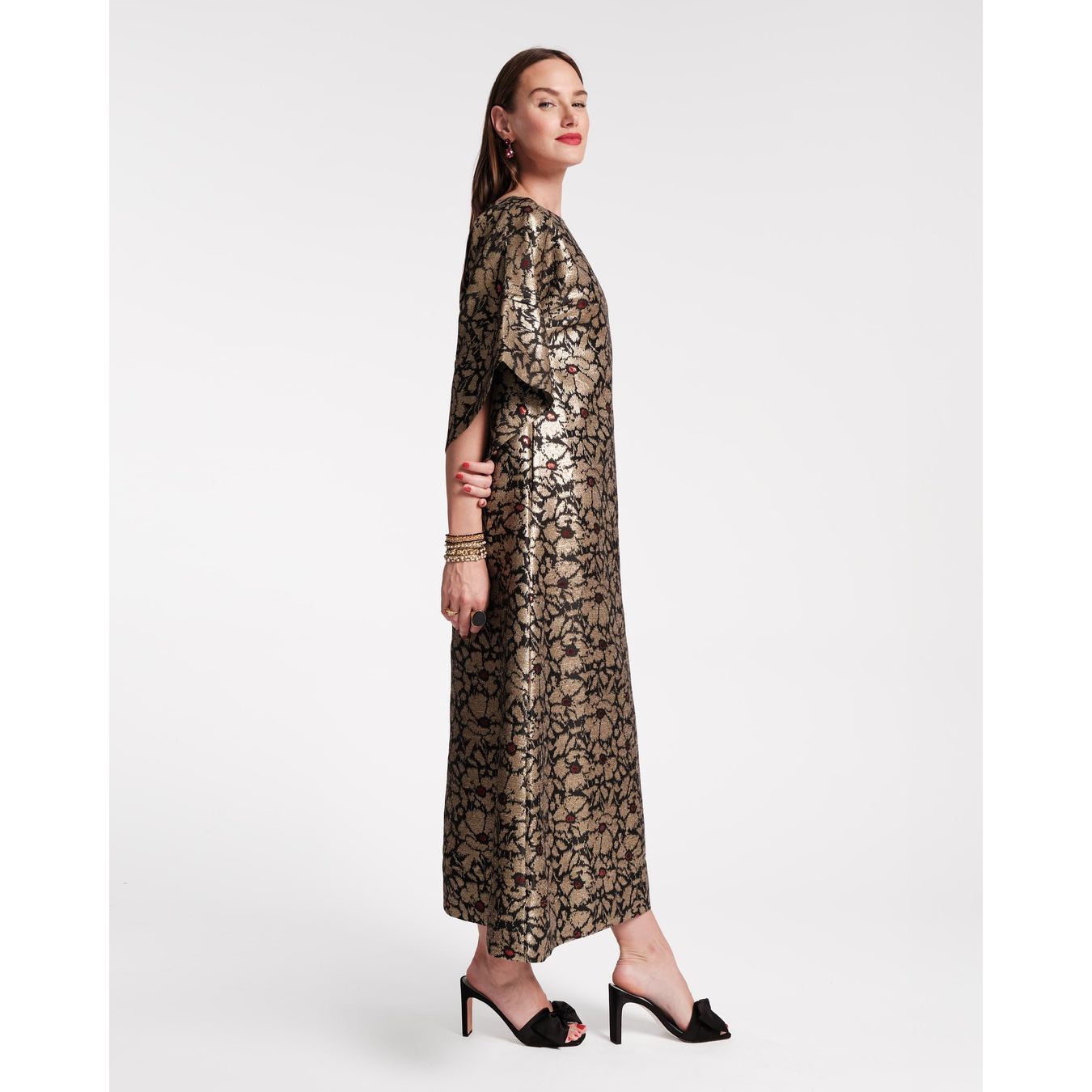 A person stands confidently in a side pose, wearing the Frances Valentine Spinnaker Maxi Dress in Winter Anemone Jacquard Black/Gold. The floor-length dress features a boatneck collar and wide sleeves and is paired with black high-heeled shoes against a plain white background.