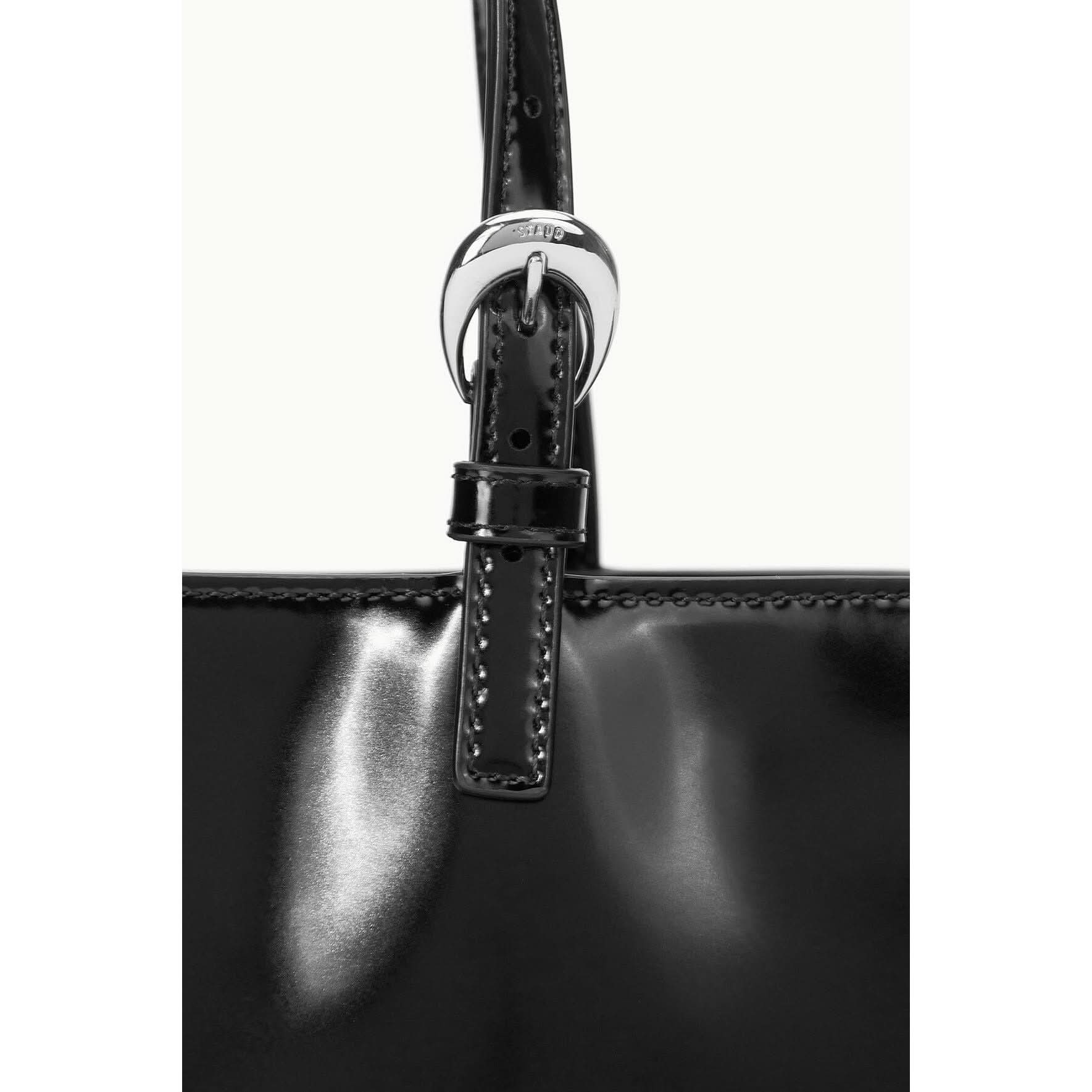 A close-up of the Staud Brando Bag Black showcases its sleek black leather design, featuring a shiny silver buckle and strap detail. The smooth texture reflects light, highlighting the craftsmanship and elegance synonymous with Staud accessories.