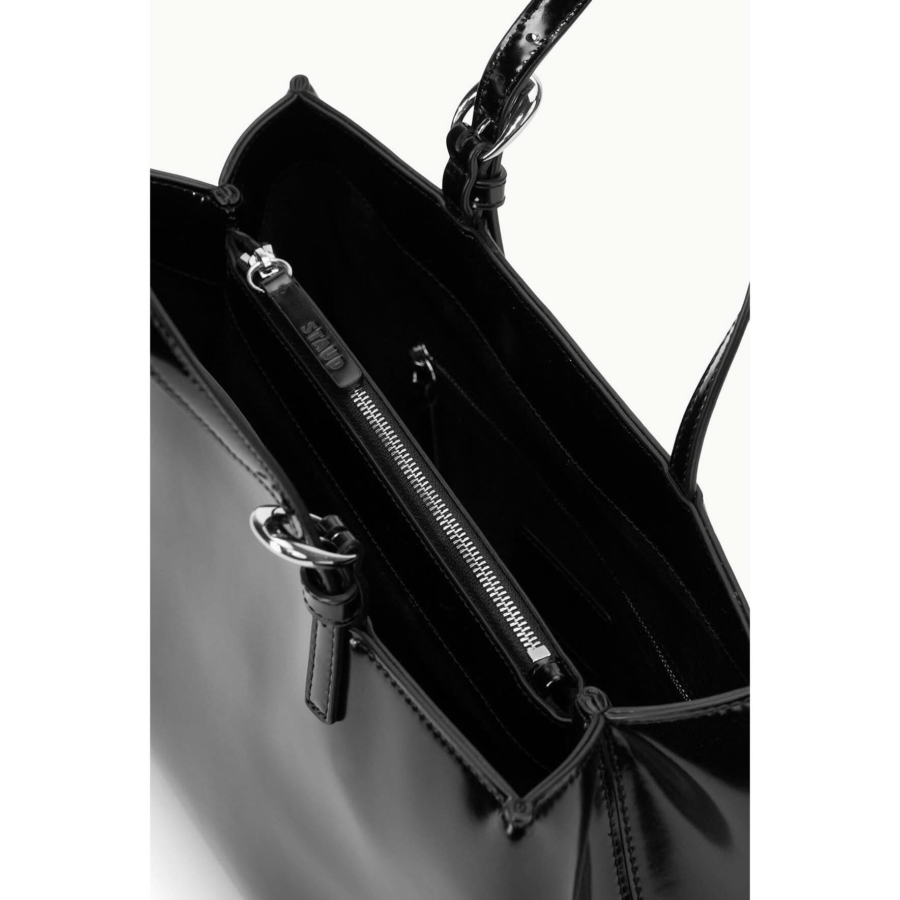 Close-up of the Staud Brando Bag in black, featuring a sleek leather design with a shiny finish. This slouchy hobo tote includes metallic rings on the handles and an inner zippered pocket labeled with Staud's brand name. The interior is lined with black fabric and has a convenient magnetic closure.