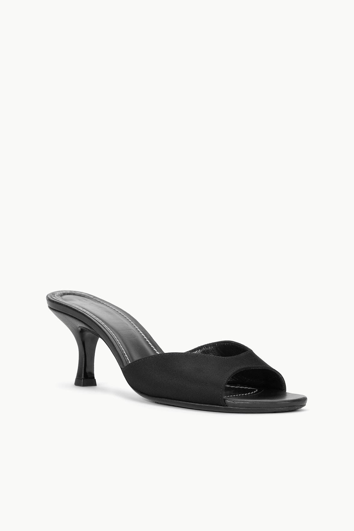 A pair of black Staud Brigitte Mules in satin with open toes and wide straps across the front, featuring a sleek, minimalist design with a slightly higher heel that's perfect for versatile dressing. The insoles have visible stitching around the edges, set against a plain, light-colored background.