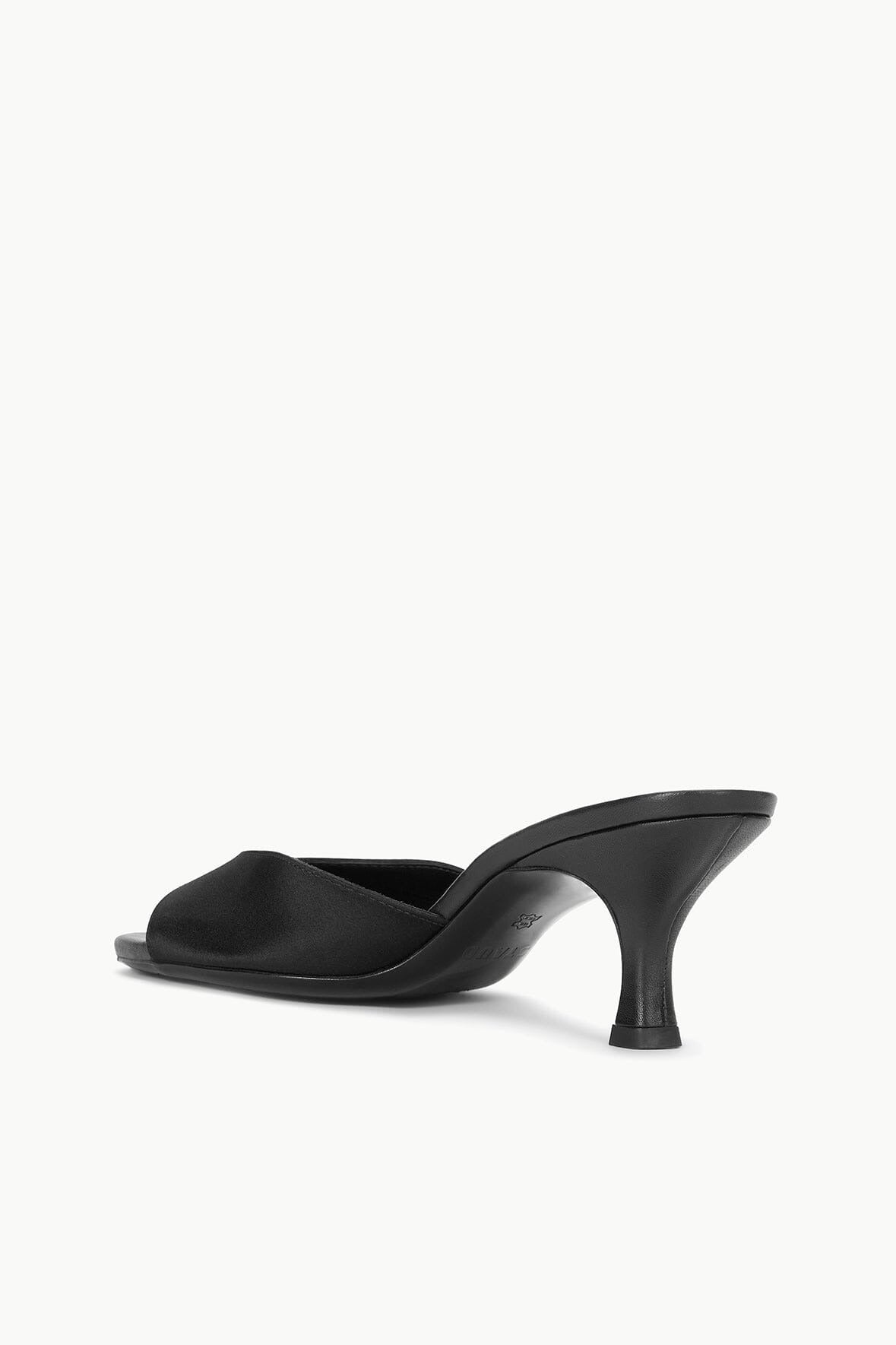 A pair of black Staud Brigitte Mules in satin with open toes and wide straps across the front, featuring a sleek, minimalist design with a slightly higher heel that's perfect for versatile dressing. The insoles have visible stitching around the edges, set against a plain, light-colored background.