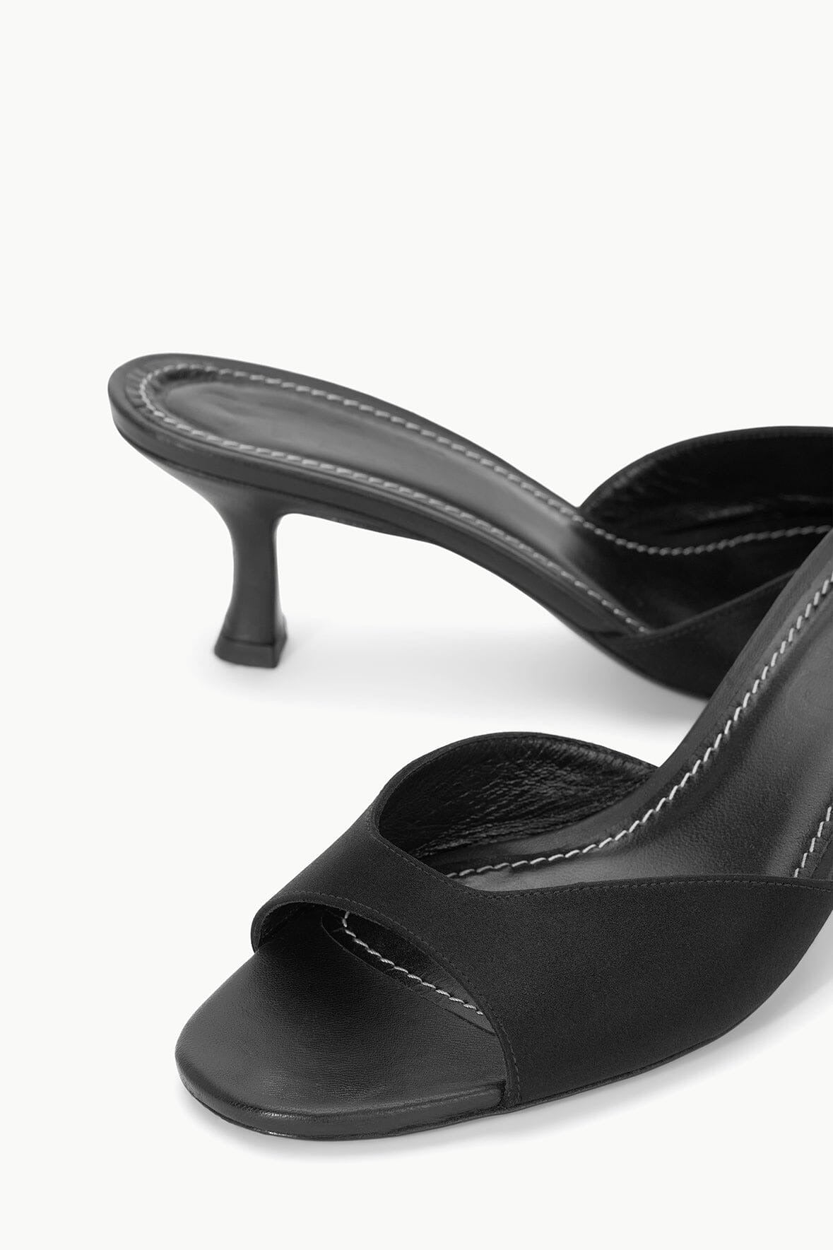 A close-up image of a pair of Staud Brigitte Mules in black satin from Staud, featuring a low, thin heel. The shoes boast a streamlined design with stitched trim along the edges, offering a sleek and elegant appearance—perfect for versatile dressing.